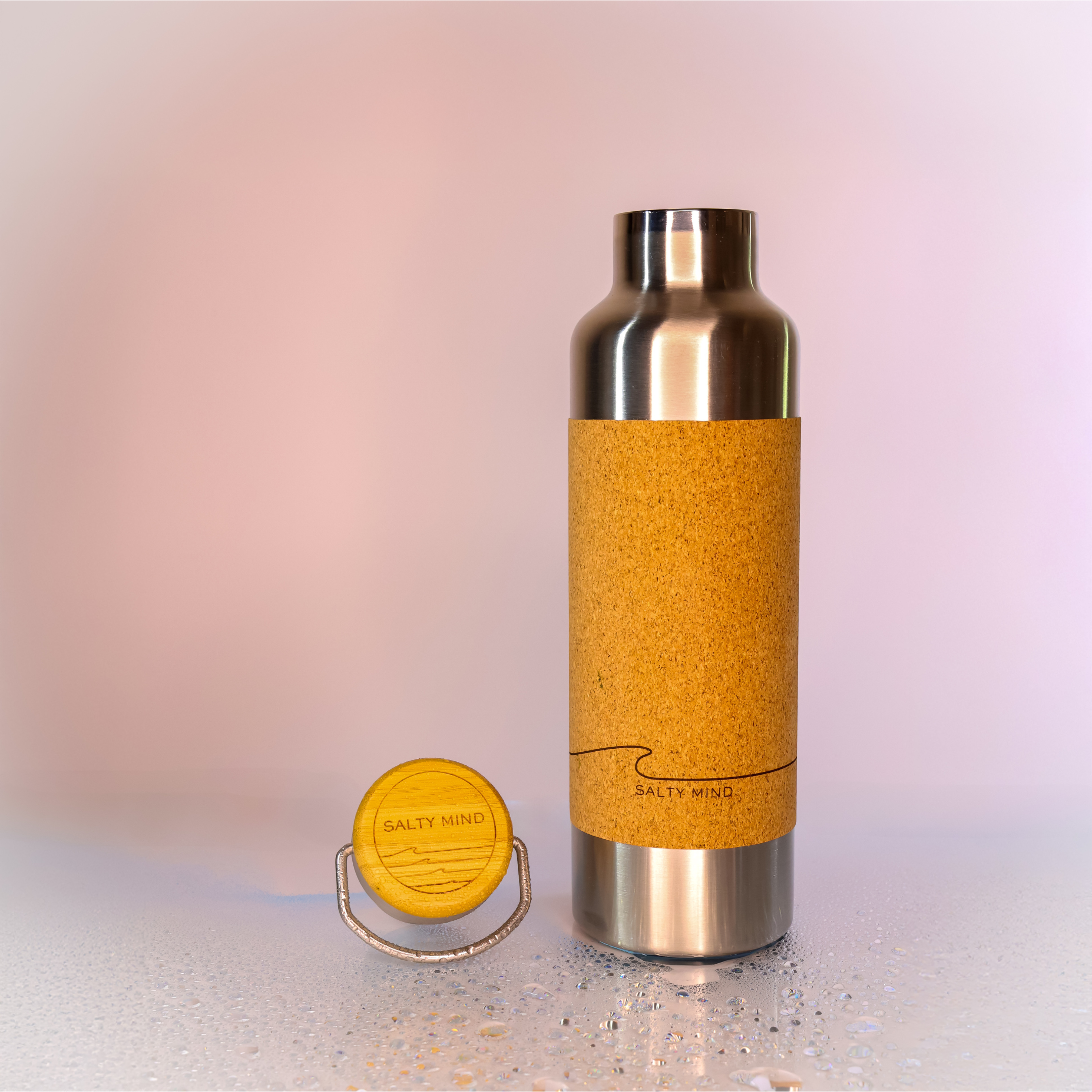 Isolierte Wasserflasche aus Edelstahl mit Korkummantelung, daneben steht der dazugehörige Deckel, auf einem nassen Untergrund.