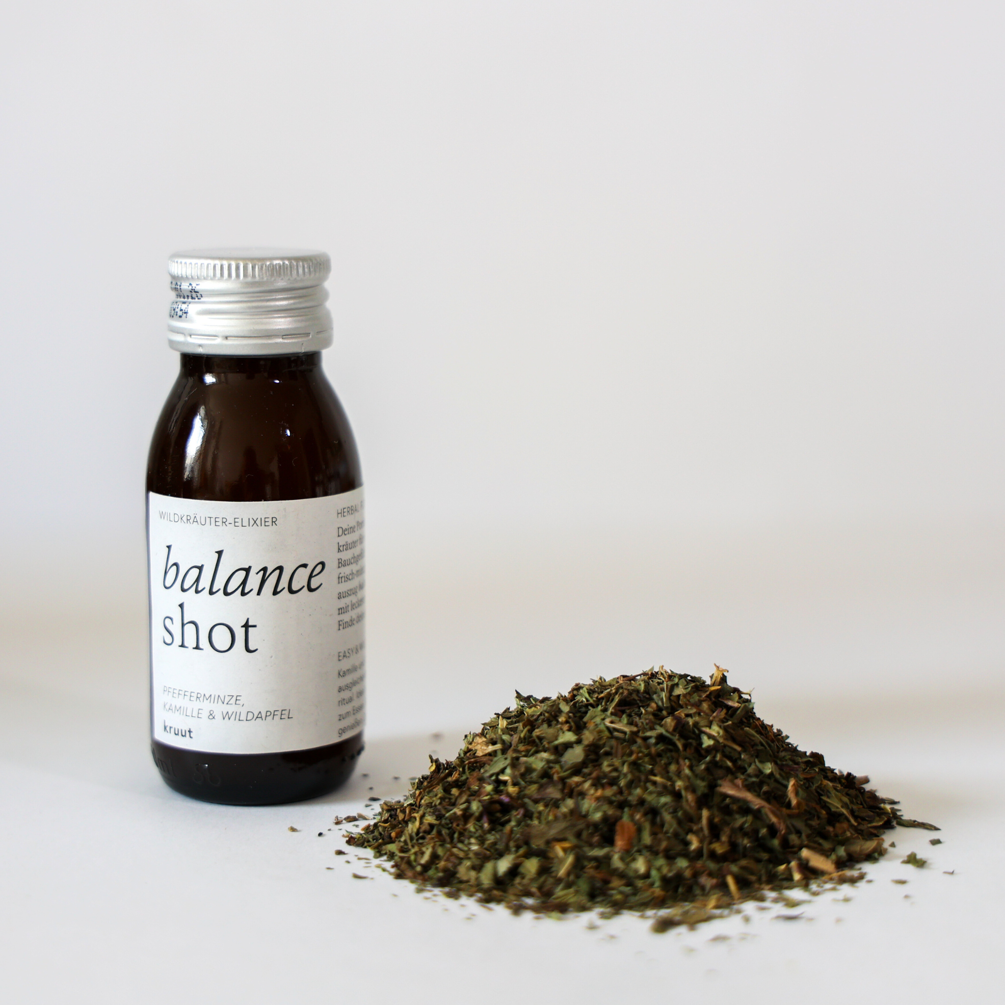 Dunkle Flasche mit der Aufschrift "balance shot" neben einem Haufen getrockneter Kräuter auf weißem Hintergrund.