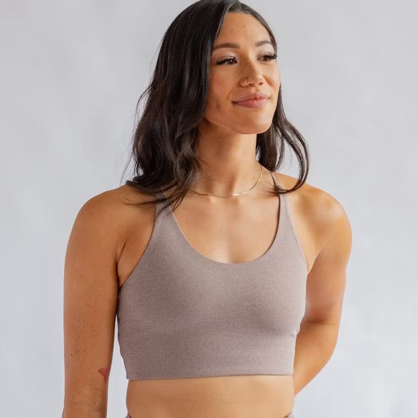 Eine Frau mit langen dunklen Haaren trägt einen hellbraunen Sport-BH und eine zarte Halskette. Sie steht vor einem schlichten hellgrauen Hintergrund und blickt mit ruhigem Blick leicht zur Seite.