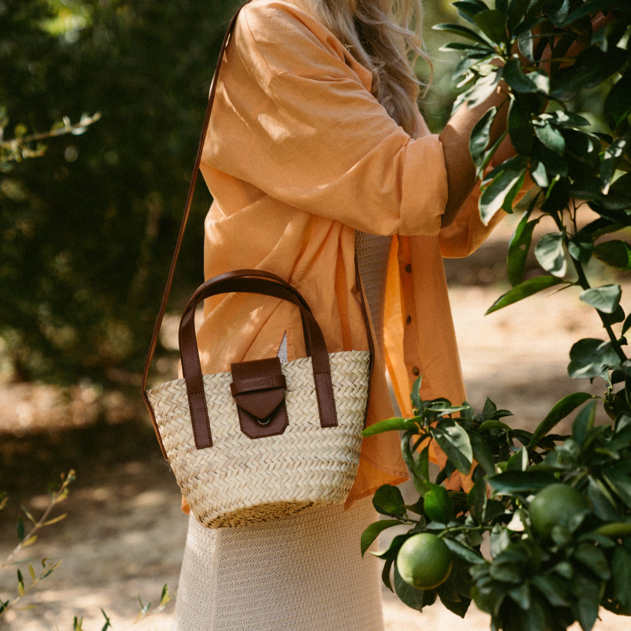 Frau in orangefarbenem Hemd, steht bei einem Baum, trägt eine Strohhandtasche mit braunen Riemen.