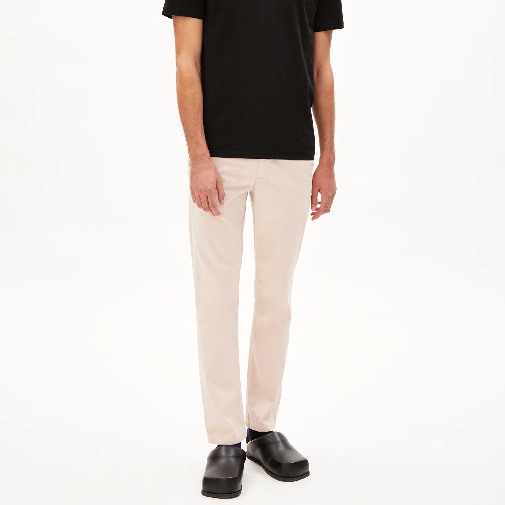 Eine Person steht vor einem schlichten weißen Hintergrund und trägt ein schwarzes kurzärmeliges T-Shirt, eine hellbeige Hose und schwarze Schuhe.