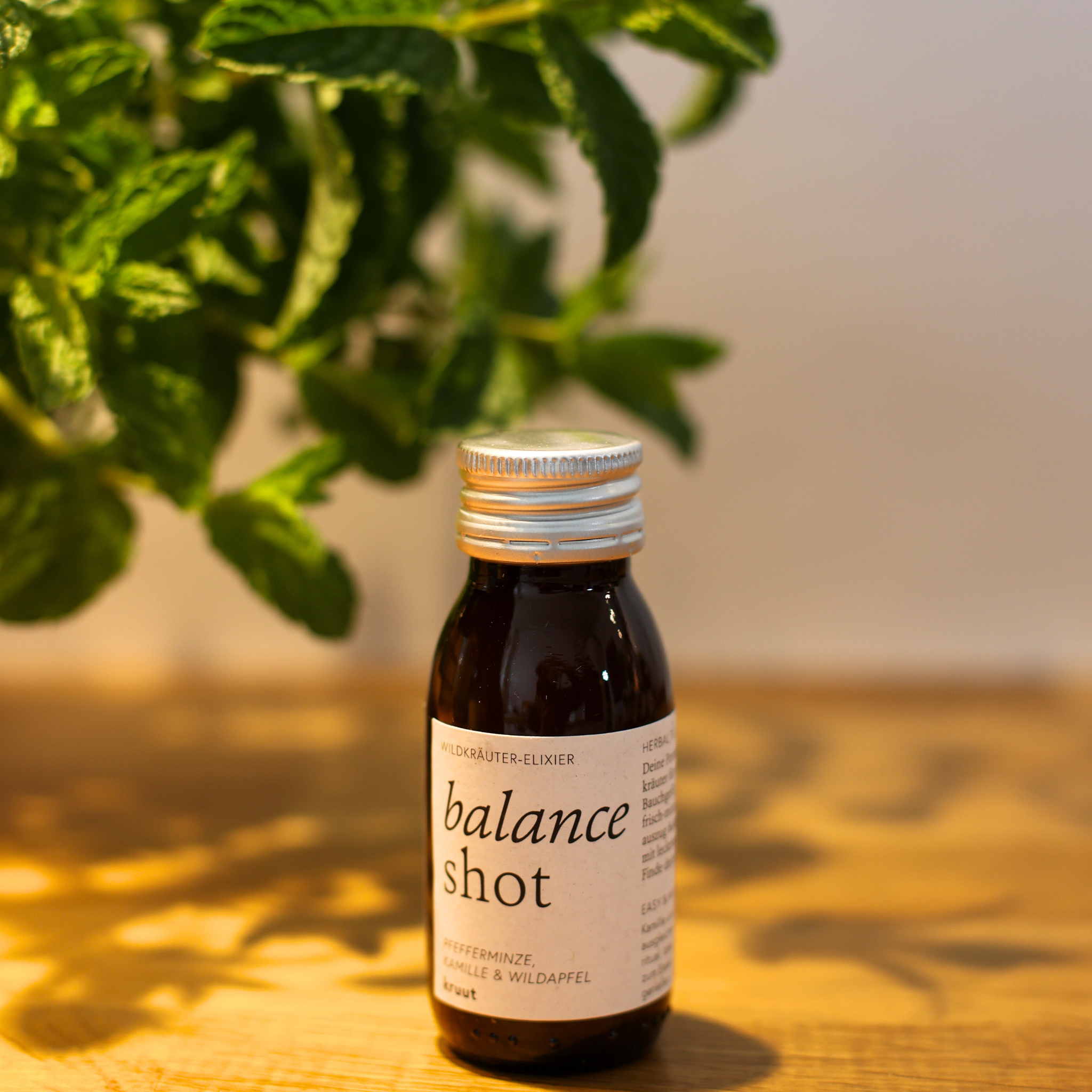 Braunes Fläschchen mit der Aufschrift "balance shot" steht auf einem Holztisch; im Hintergrund grüne Blätter.