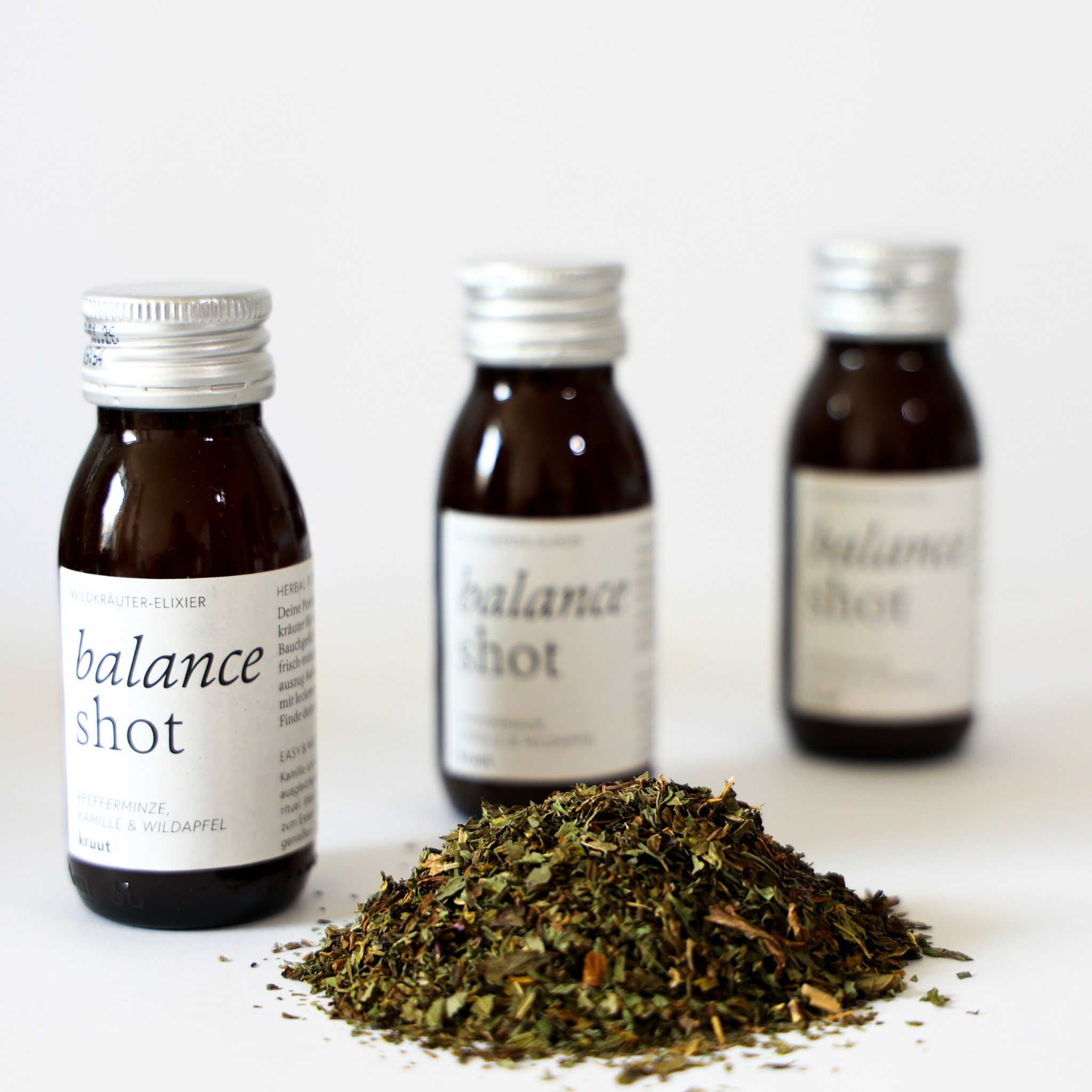 Drei braune Flaschen mit "balance shot"-Etikett, im Vordergrund ein Haufen getrockneter Kräuter auf weißem Hintergrund.
