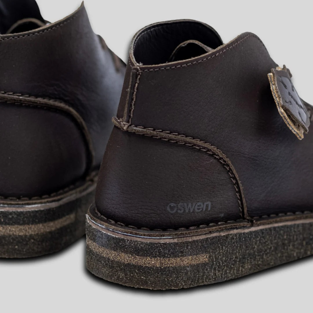 Nahaufnahme der Rück- und Seitenansicht von zwei dunkelbraunen Lederschuhen mit sichtbaren Nähten, strukturierten Sohlen und dem Wort "Oswen" auf dem Absatz. Der Hintergrund ist einfarbig hellgrau.