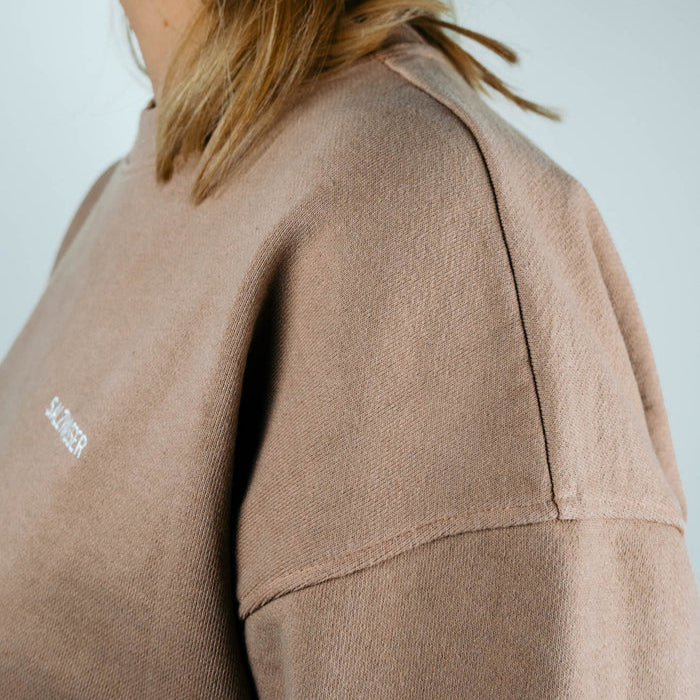 Nahaufnahme einer Person, die ein hellbraunes Sweatshirt mit sichtbaren Nähten an der Schulter trägt. Ein Teil der blonden Haare der Person ist sichtbar, und nur der Oberkörper und der Hals sind zu sehen. Der Hintergrund ist einfarbig und hell gefärbt.