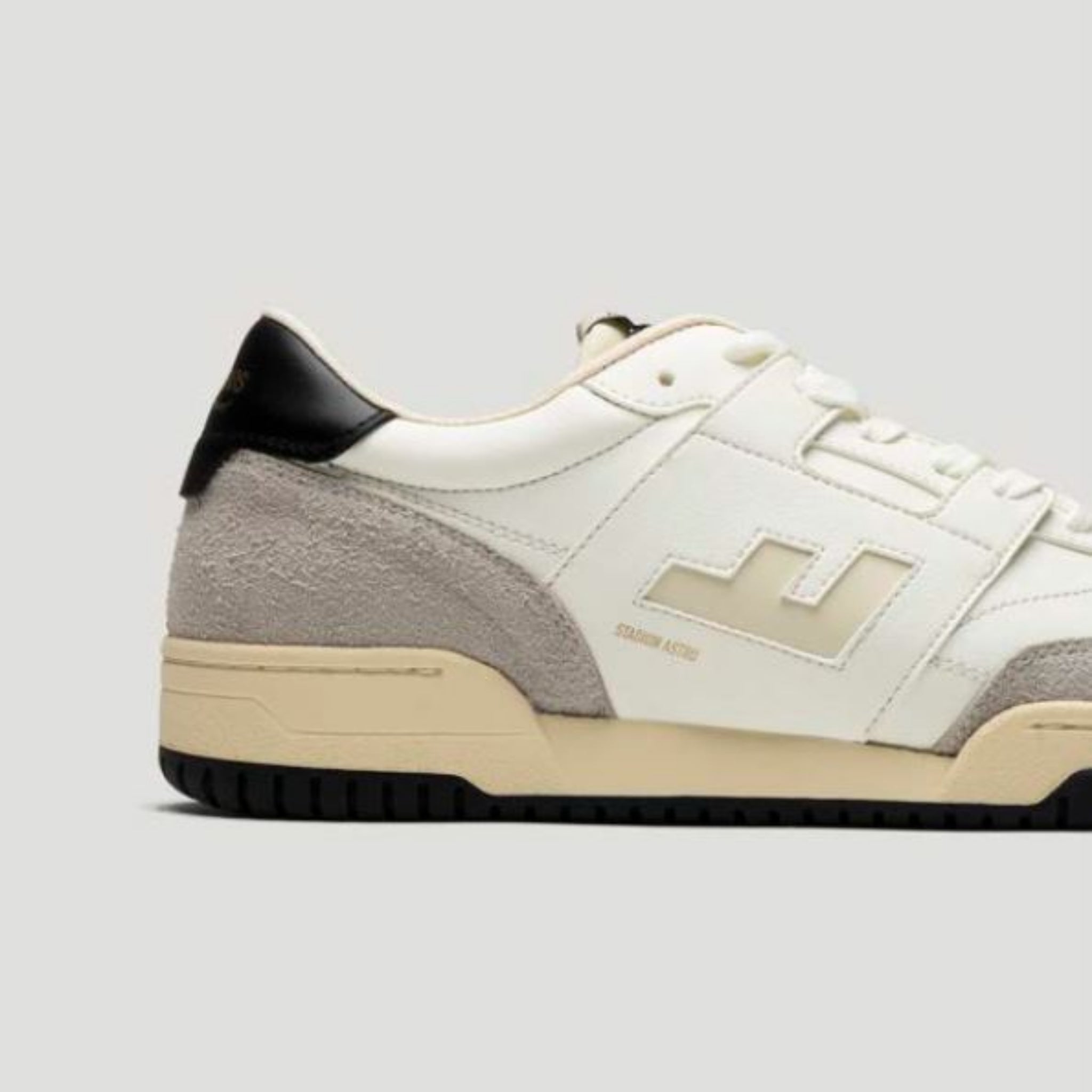 Ein weißer Ledersneaker mit hellgrauem Wildlederabsatz und beiger Sohle, mit einem erhabenen geometrischen Logo an der Seite und einer schwarzen Fersenlasche, die vor einem schlichten hellgrauen Hintergrund gezeigt wird.