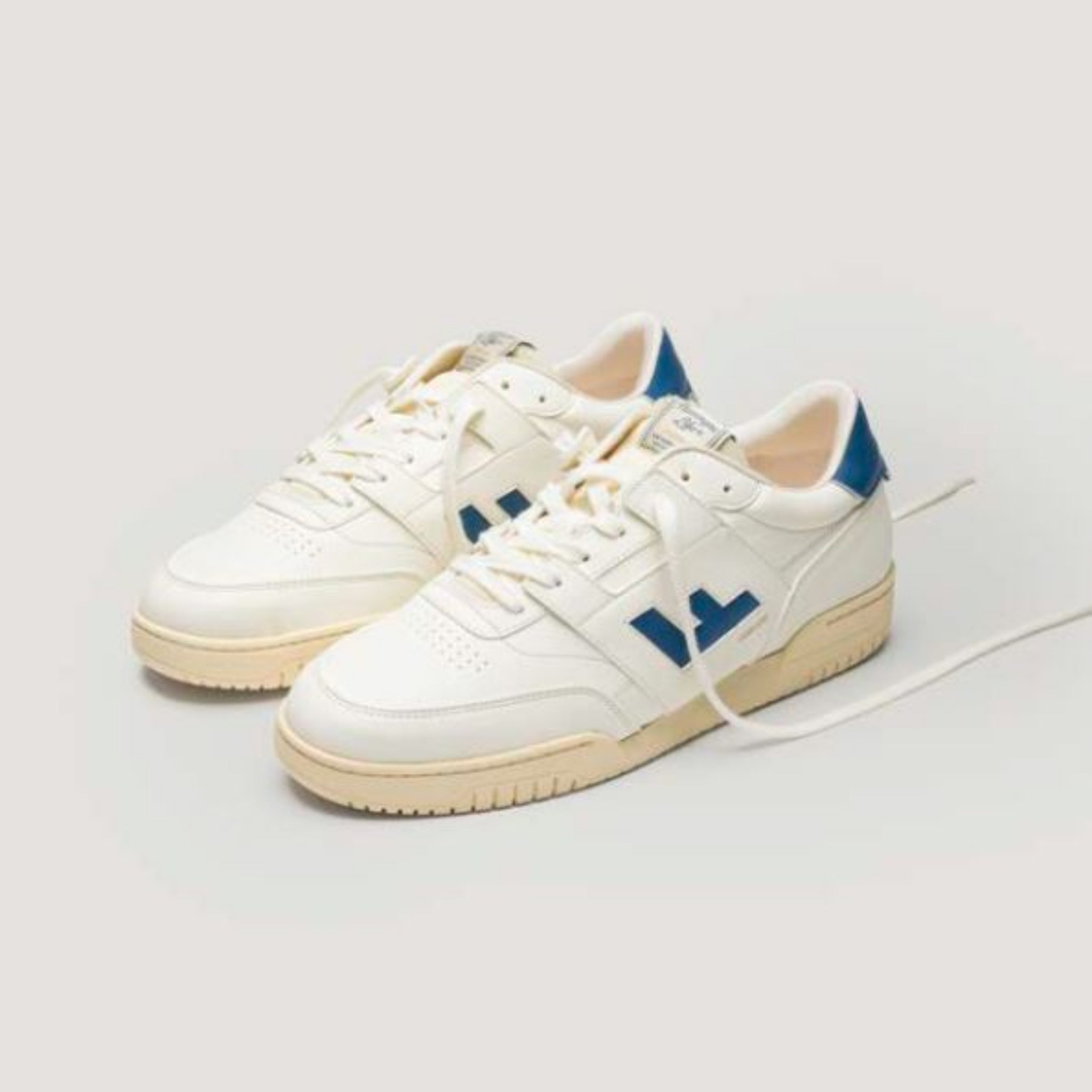 Ein Paar weißer Low-Top-Sneaker mit cremefarbener Sohle und blauen Akzenten an der Ferse und an der Seite, dargestellt auf hellem Hintergrund mit einem teilweise geöffneten Schnürsenkel.