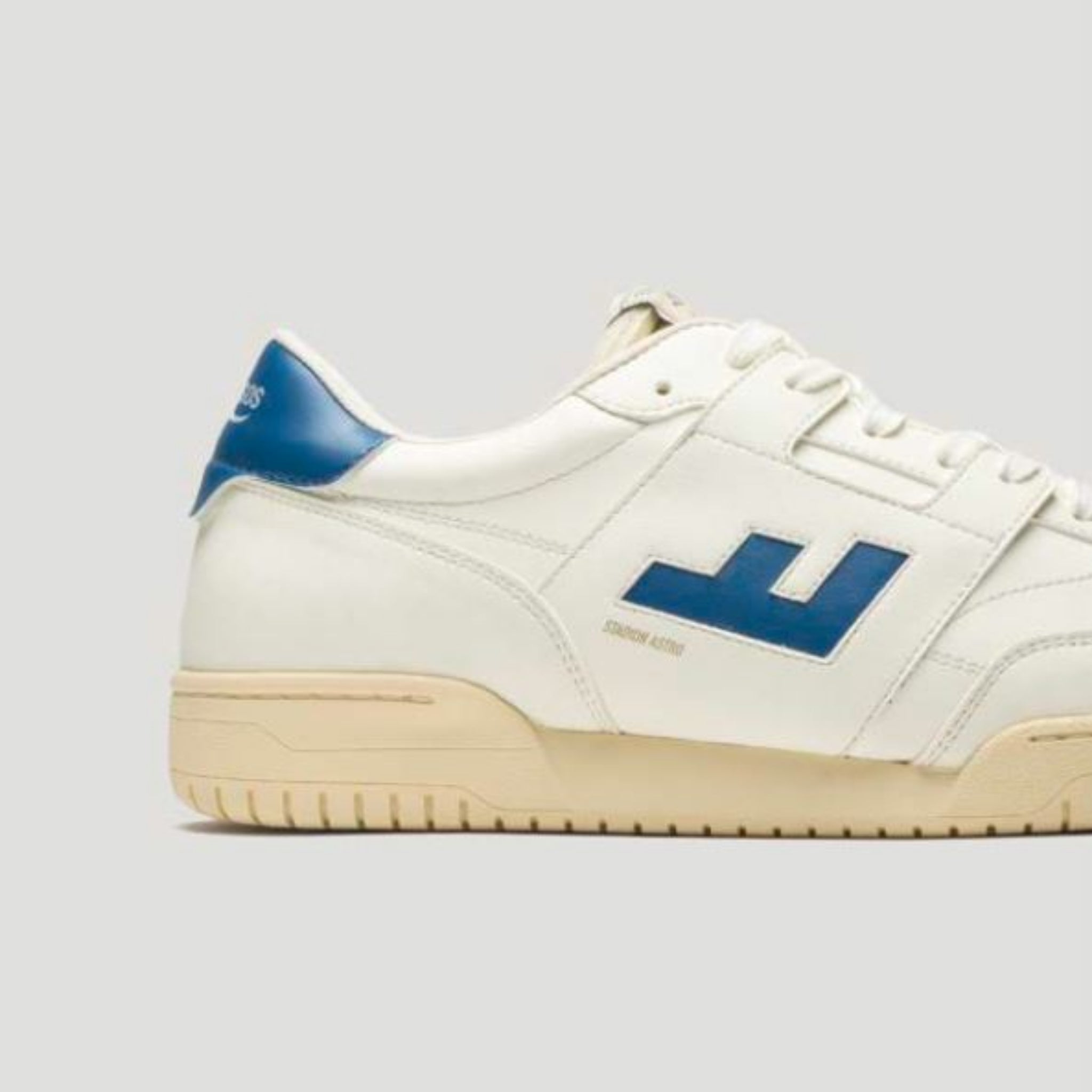 Ein weißer Sneaker mit einem blauen L-förmigen Logo an der Seite, einem blauen Fersendetail, einer beigen Sohle und weißen Schnürsenkeln, dargestellt auf einem hellgrauen Hintergrund.
