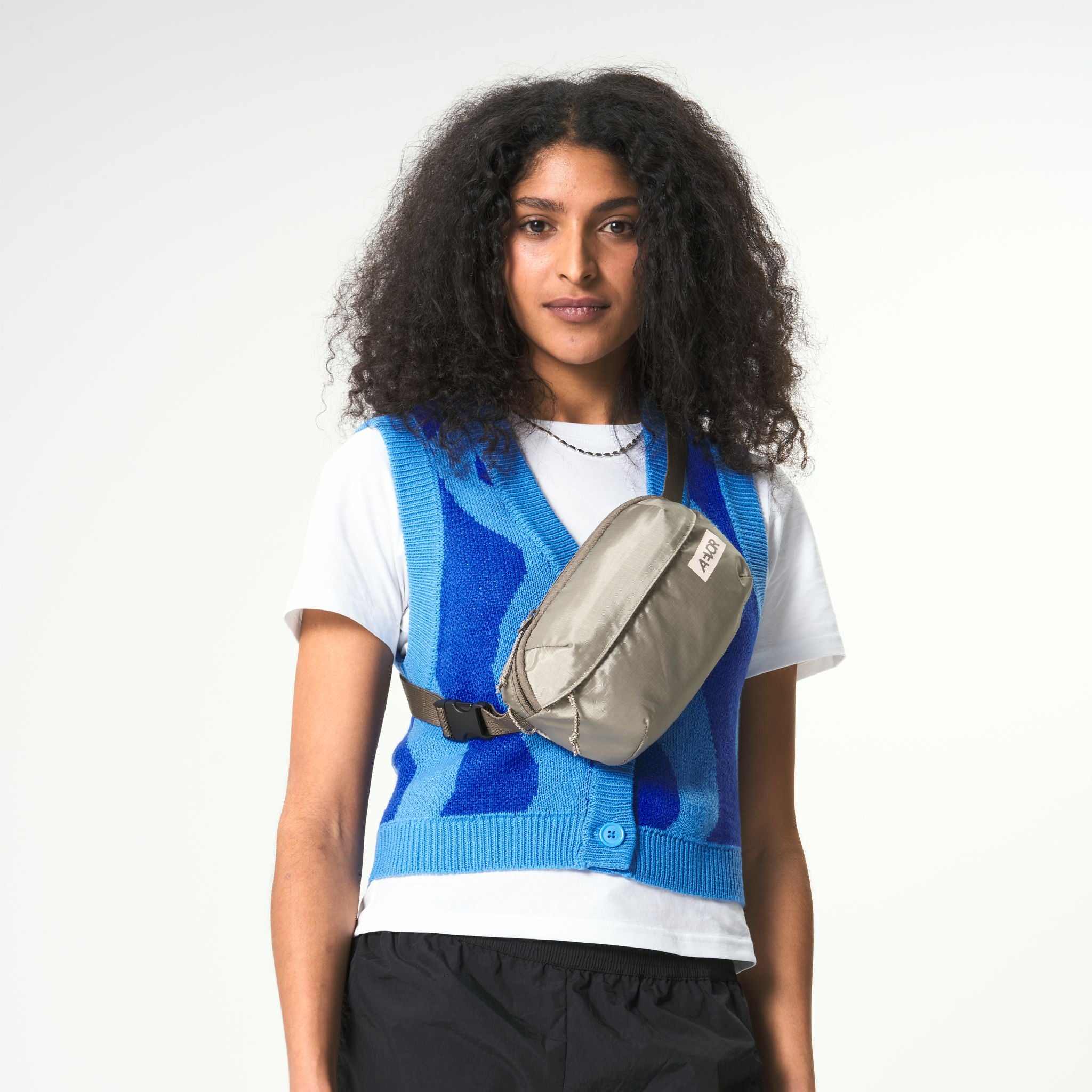 Person mit lockigem Haar trägt einen blauen Strickwesten über einem weißen T-Shirt und eine graue Umhängetasche.
