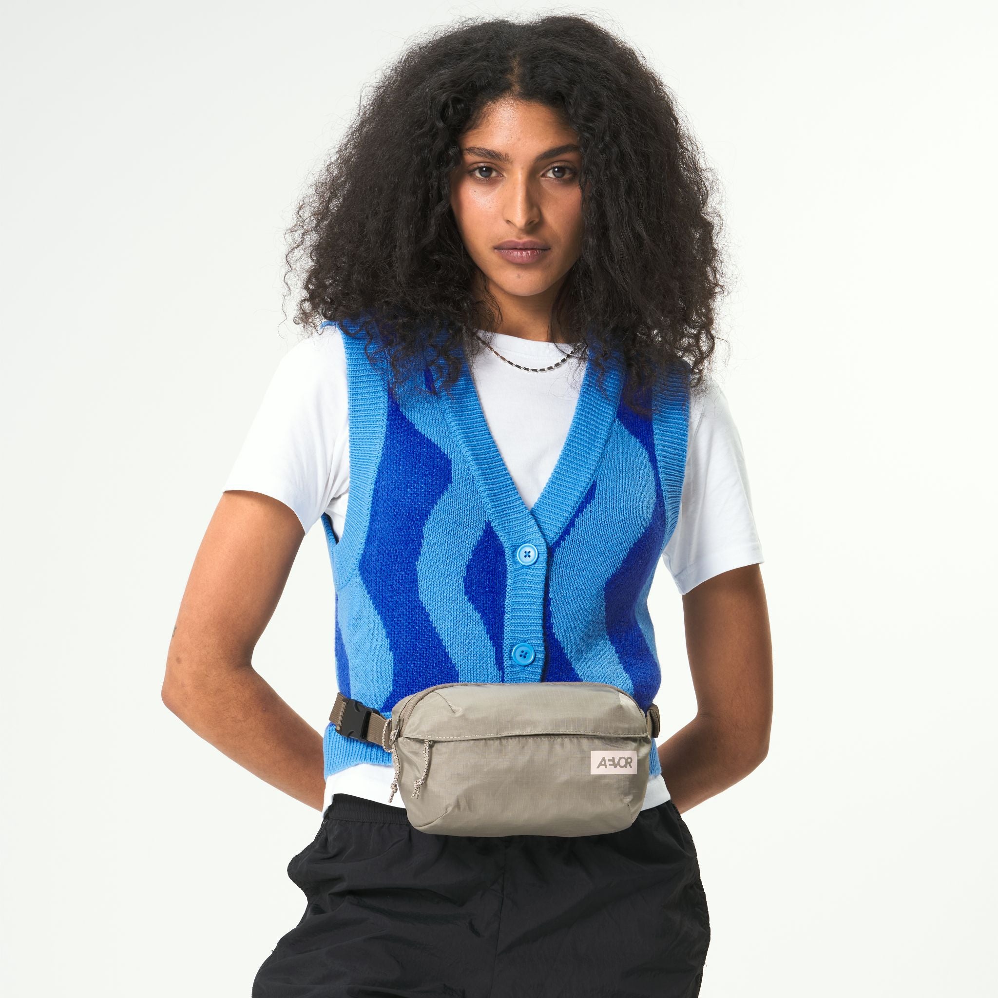 Person mit lockigem Haar trägt eine blaue Weste über einem weißen T-Shirt und eine beige Bauchtasche, posiert vor weißem Hintergrund.