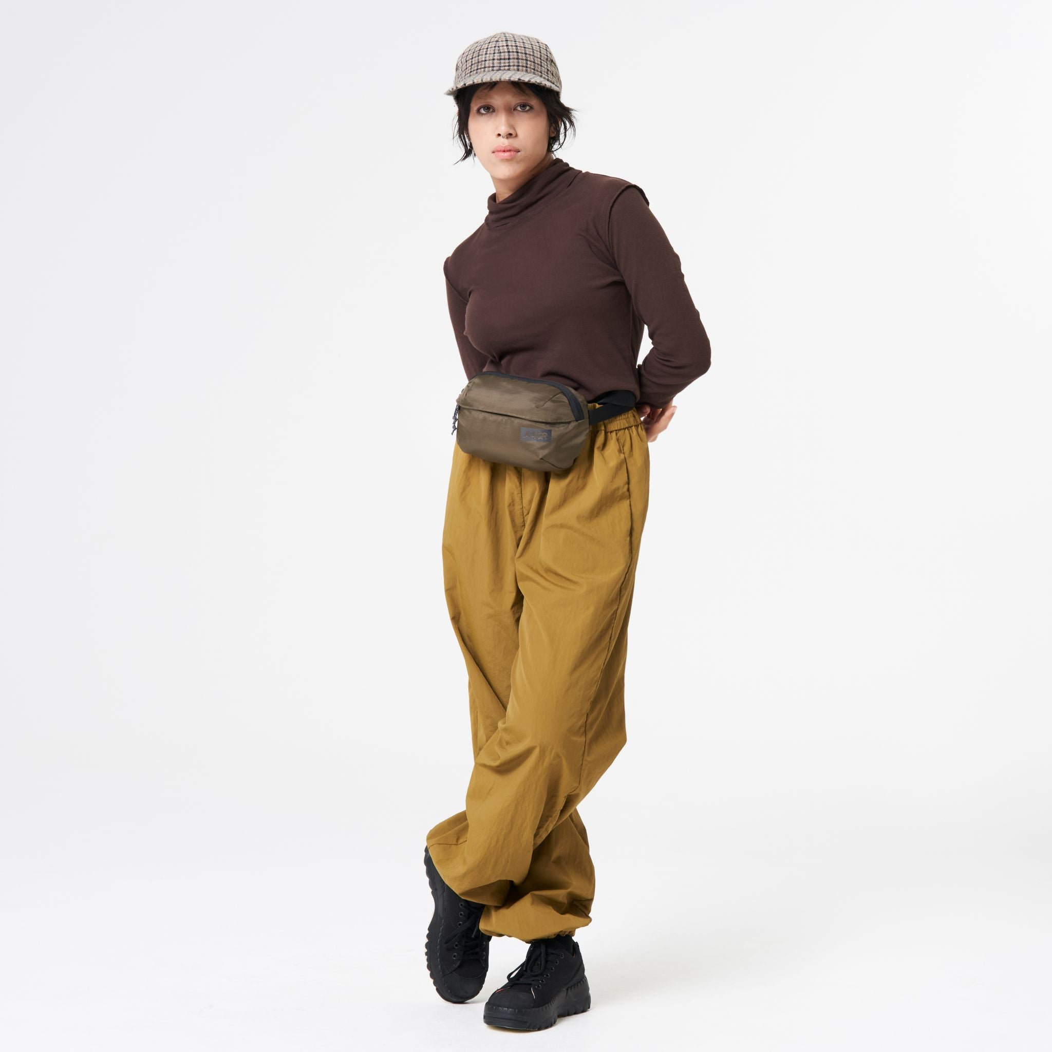 Person in a brown shirt, mustard pants, checkered cap, and belt bag poses in a studio.