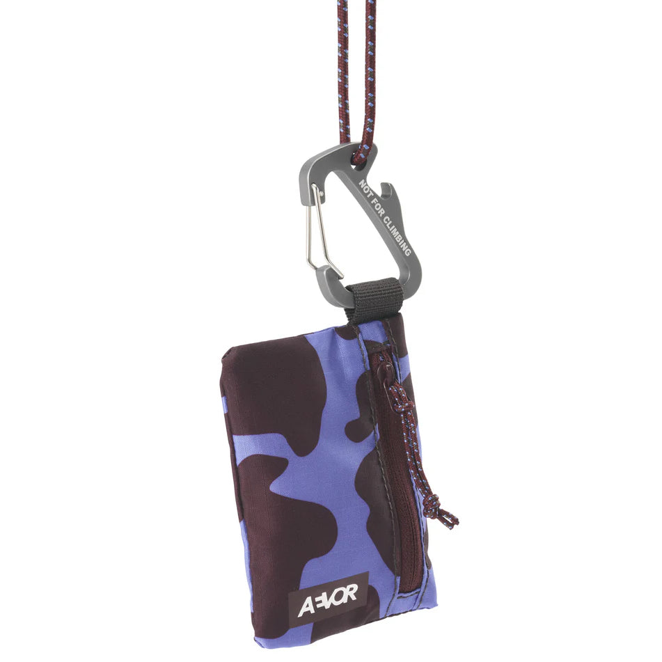 Eine kleine Reißverschlusstasche mit blauem und dunkelbraunem Tarnmuster, befestigt an einem Seil und einem silbernen Karabiner. Auf der Vorderseite des Beutels steht AVOR geschrieben.