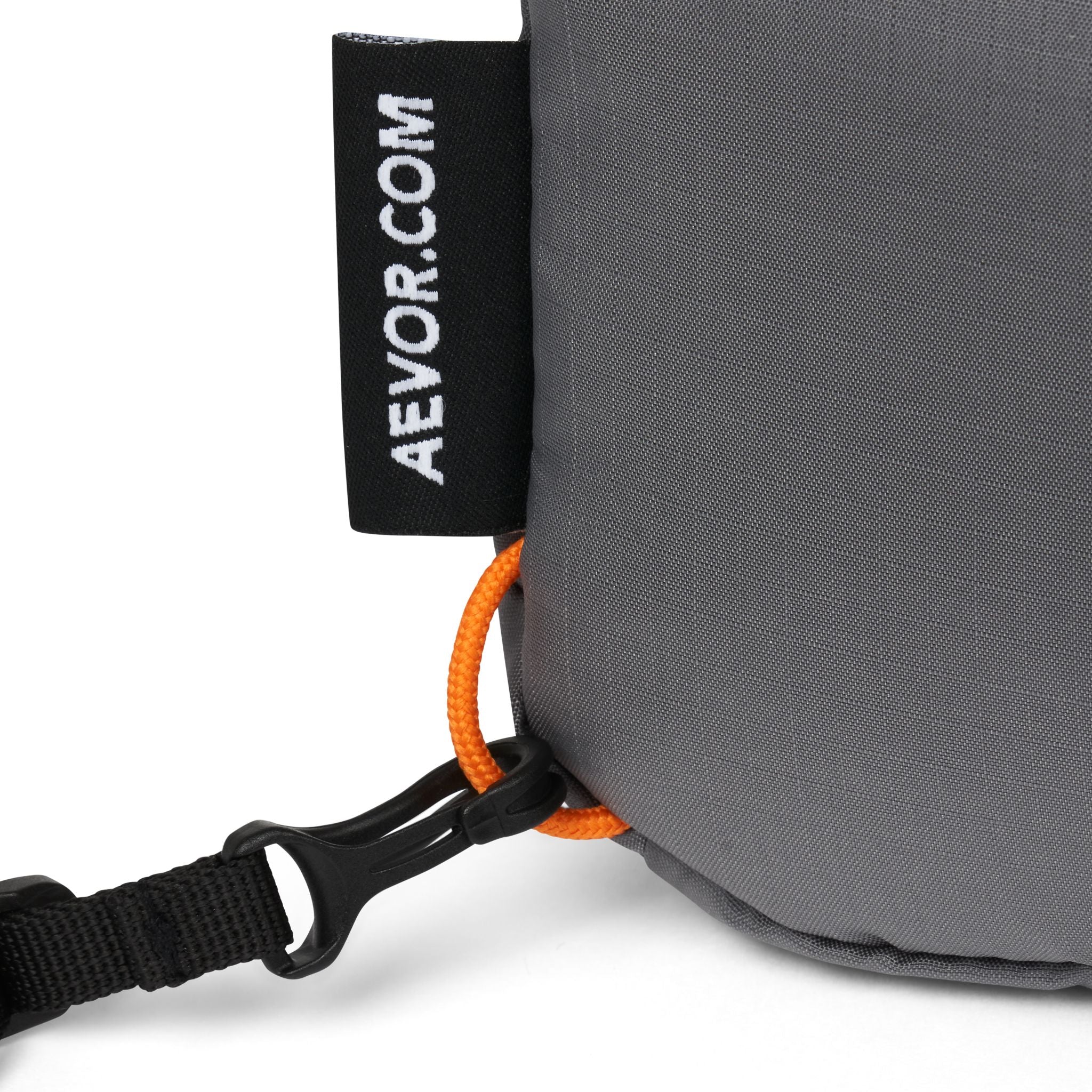 Nahaufnahme eines grauen Rucksacks mit einem schwarzen Etikett "AEVOR.COM" und einem orangefarbenen Band mit schwarzem Clip.