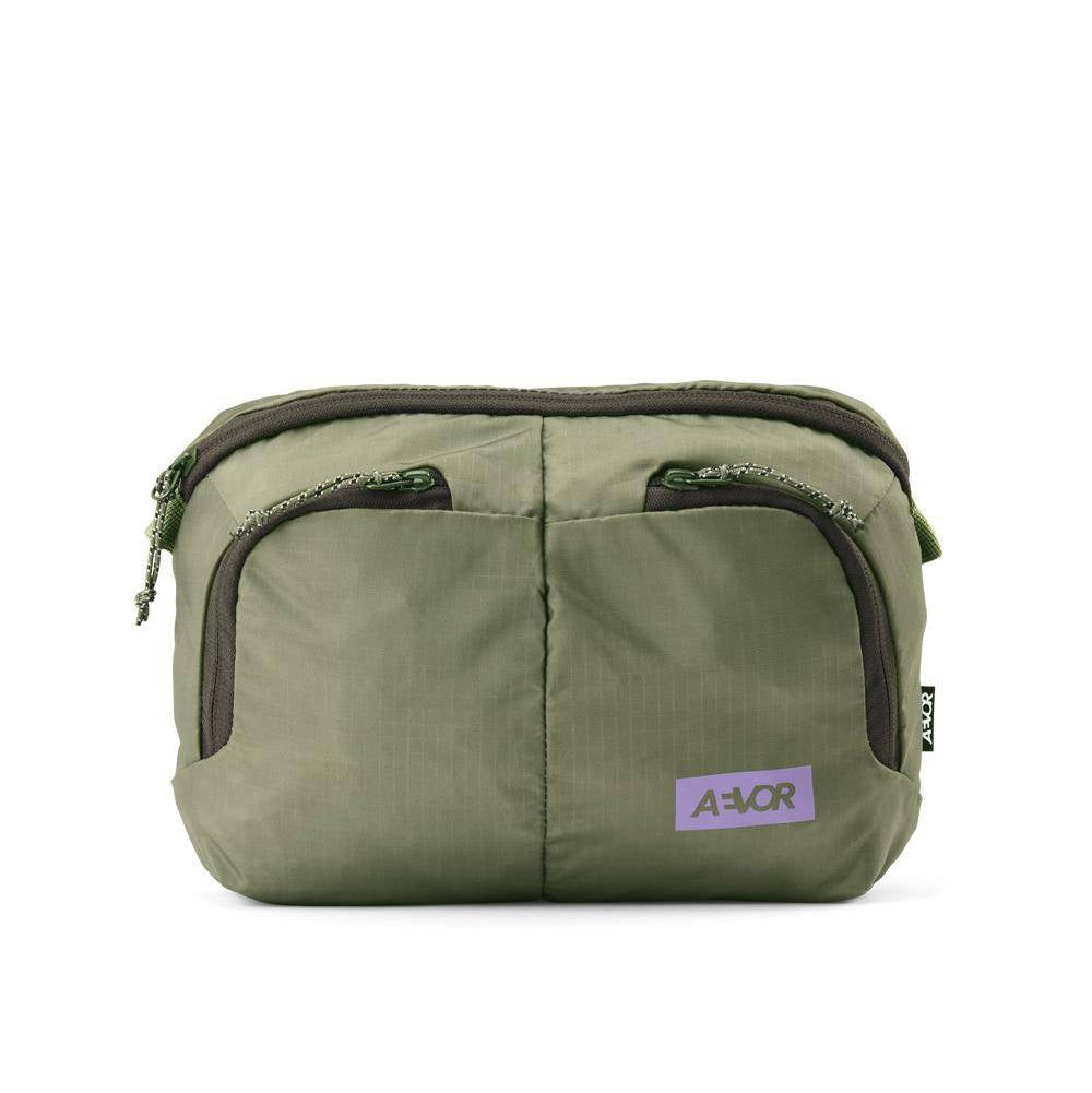 Eine kleine, olivgrüne Umhängetasche mit zwei Reißverschlusstaschen auf der Vorderseite, einem Reißverschluss oben und einem Logoaufnäher mit der Aufschrift A-VOR auf der Vorderseite. Der Stoff hat ein leicht strukturiertes Aussehen.