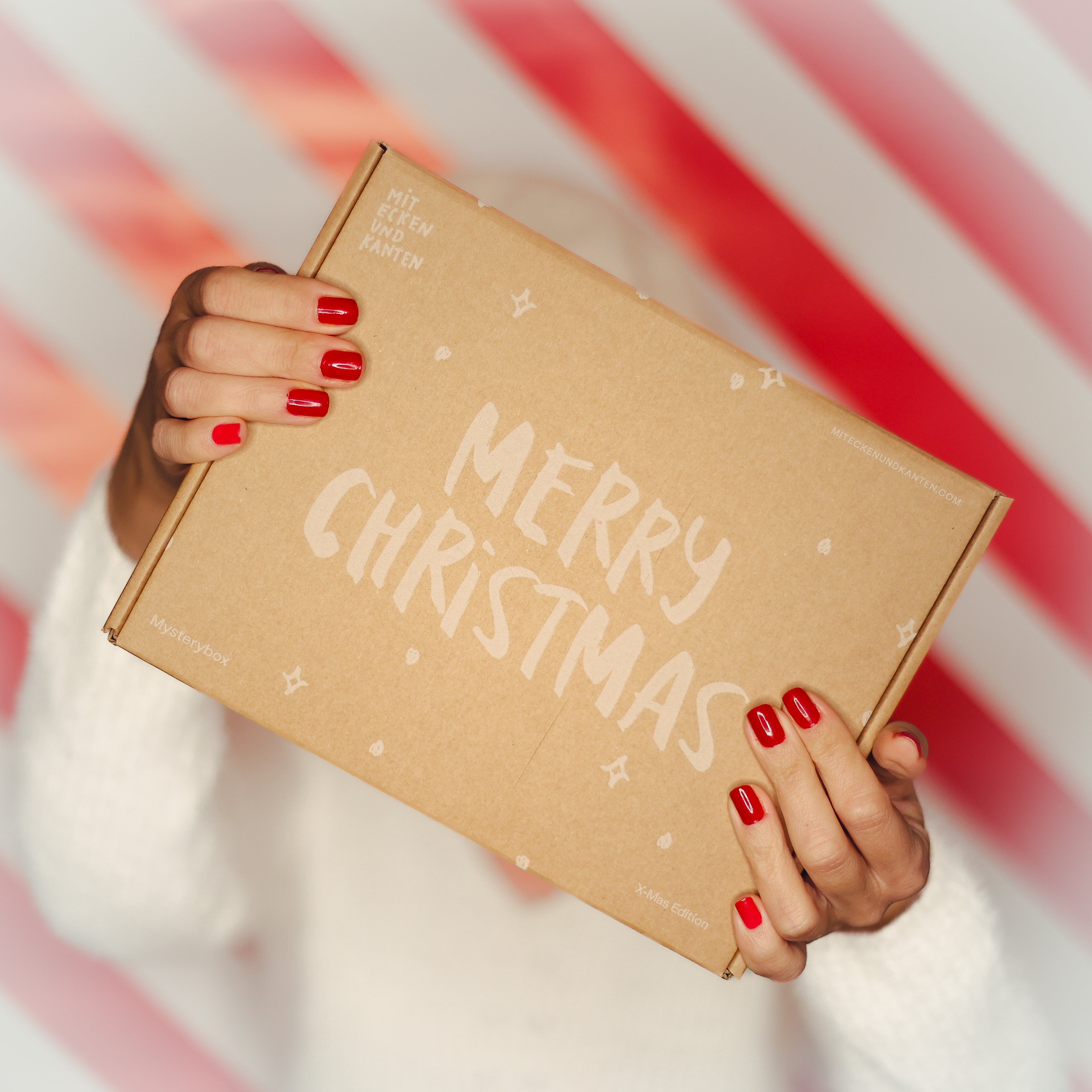Eine Person hält eine Box mit der Aufschrift "Merry Christmas" vor einem rot-weiß gestreiften Hintergrund.