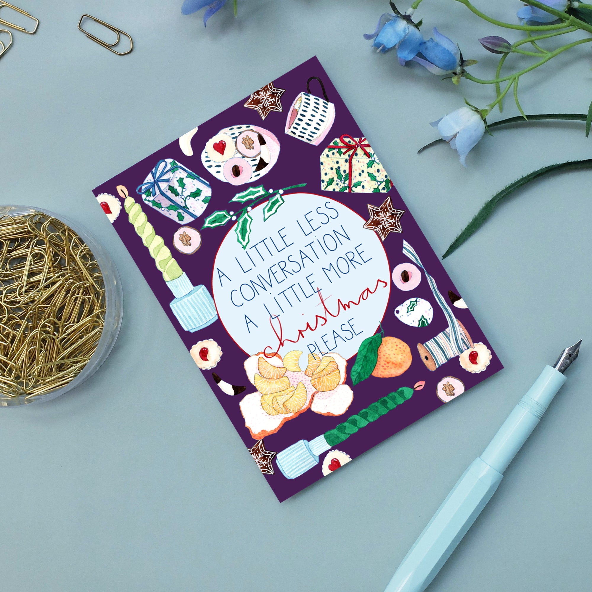 Eine farbenfrohe Feiertagskarte auf einem Schreibtisch trägt die Aufschrift "Etwas weniger Konversation, etwas mehr Weihnachten bitte", umgeben von festlichen Lebensmittelillustrationen. In der Nähe befinden sich eine Dose mit Büroklammern, ein blauer Stift und blaue Blumen.