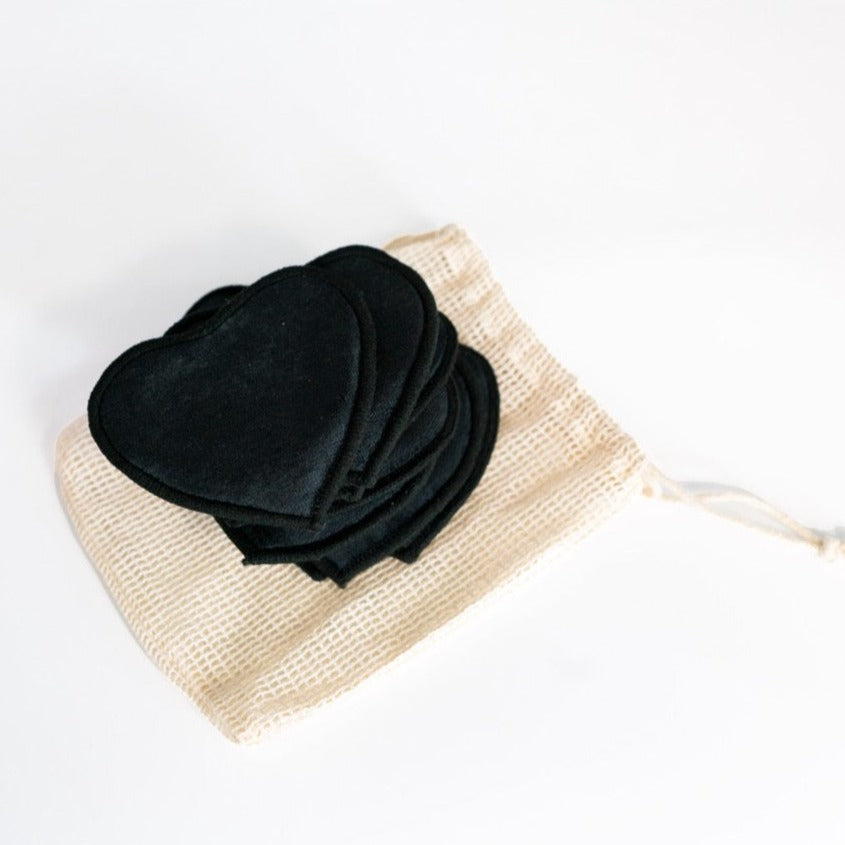 Schwarze, herzförmige Stoffpads liegen auf einem beigen Netzbeutel auf einem weißen Hintergrund.
