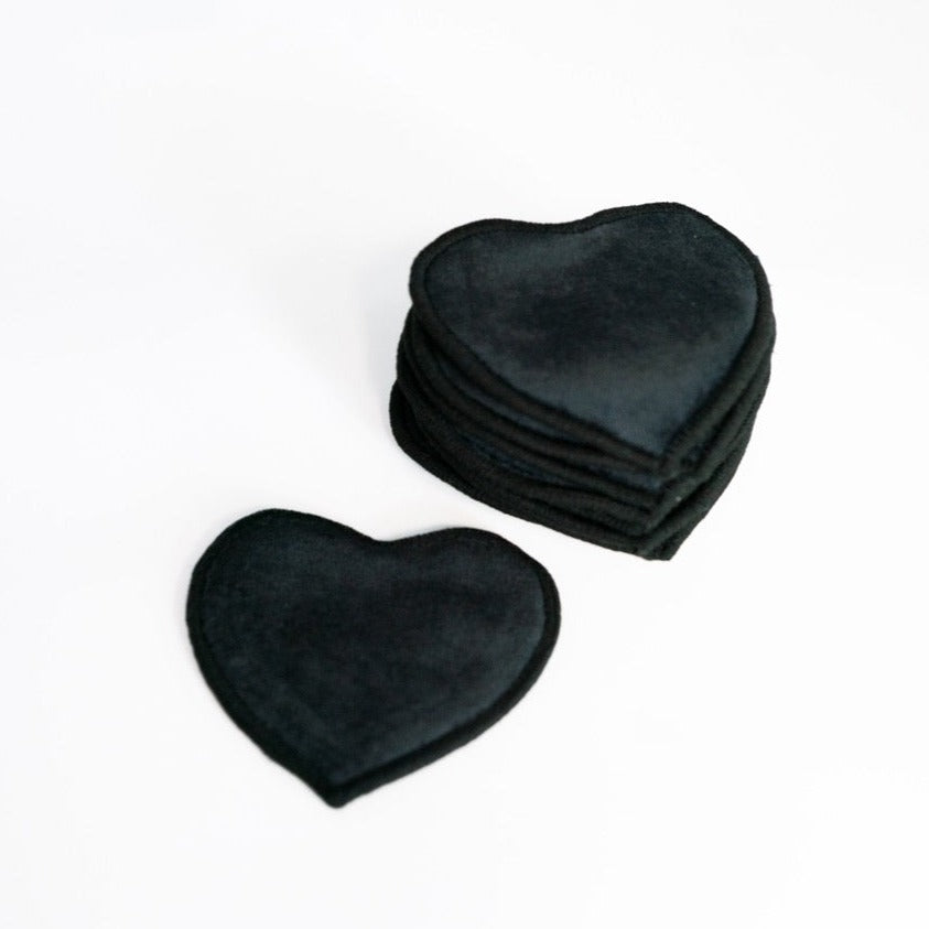 Ein Stapel schwarzer herzförmiger Stoffpads liegt auf einem weißen Hintergrund, daneben ein einzelnes Pad.