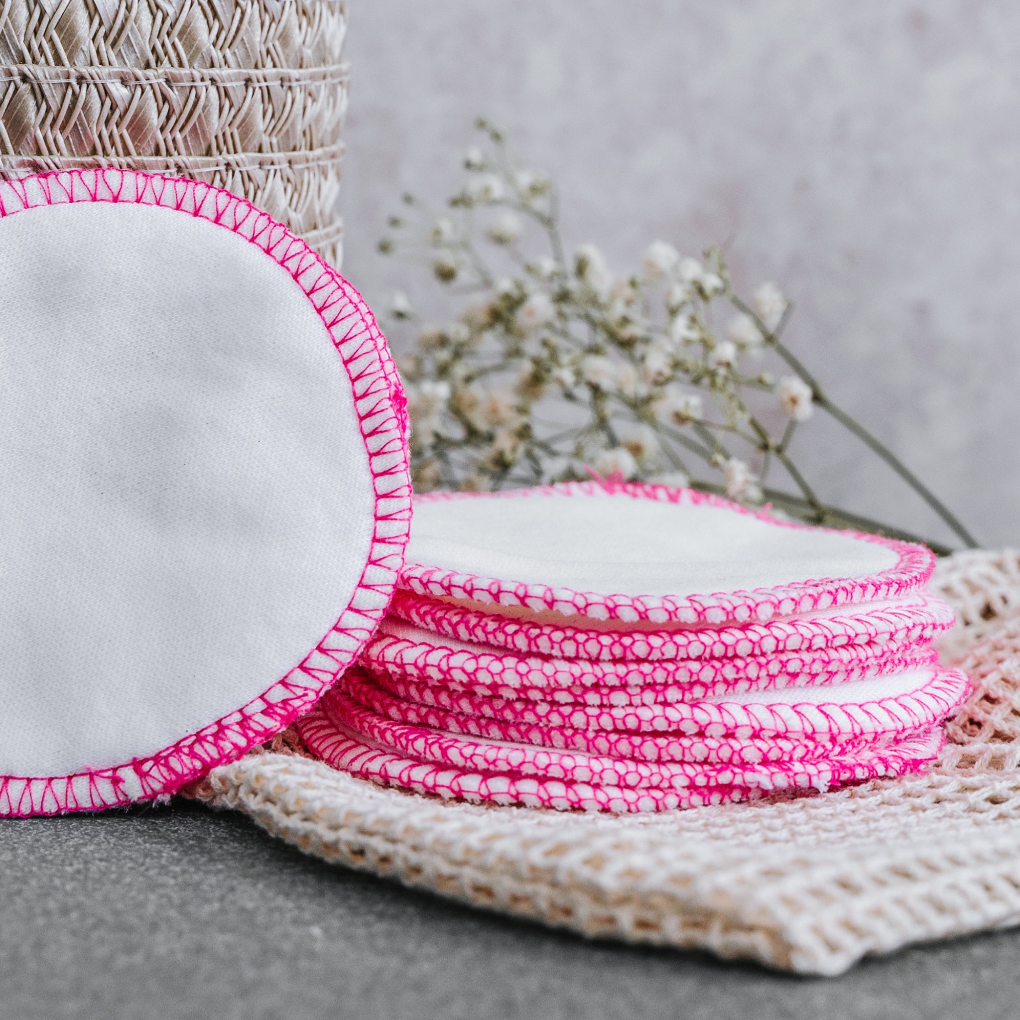 Stapel wiederverwendbarer Abschminkpads mit rosa Rändern vor einem Korb und Blumen im Hintergrund.