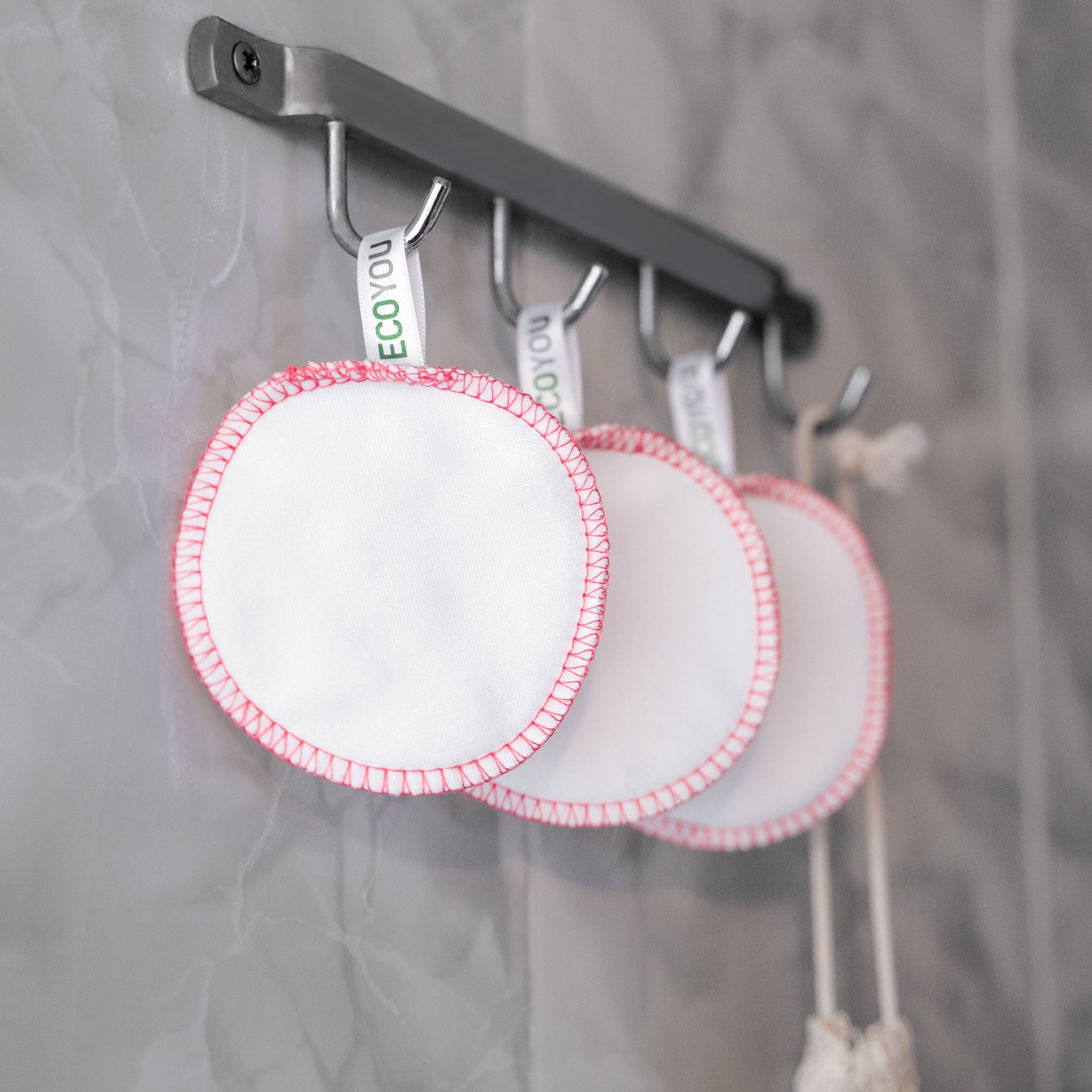 Drei weiße, wiederverwendbare Wattepads mit rotem Rand hängen an Haken vor einer grauen Wand.