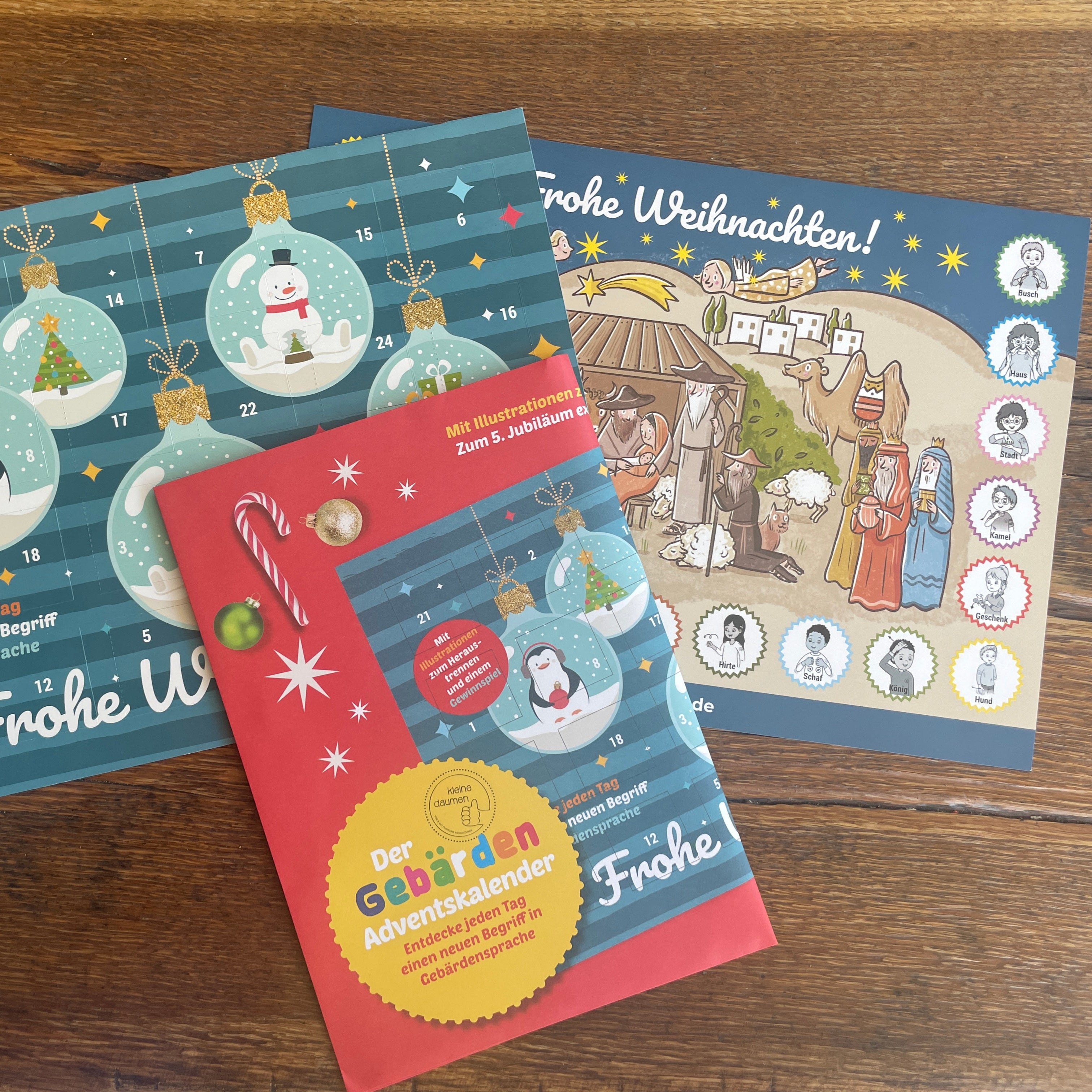 Ein Set aus farbenfrohen deutschen Adventskalendern und einem Heft mit weihnachtlichen Motiven, die Illustrationen von Pinguinen, Ornamenten, einer Krippe und festlichen Dekorationen zeigen, die alle auf einer hölzernen Unterlage ausgelegt sind.