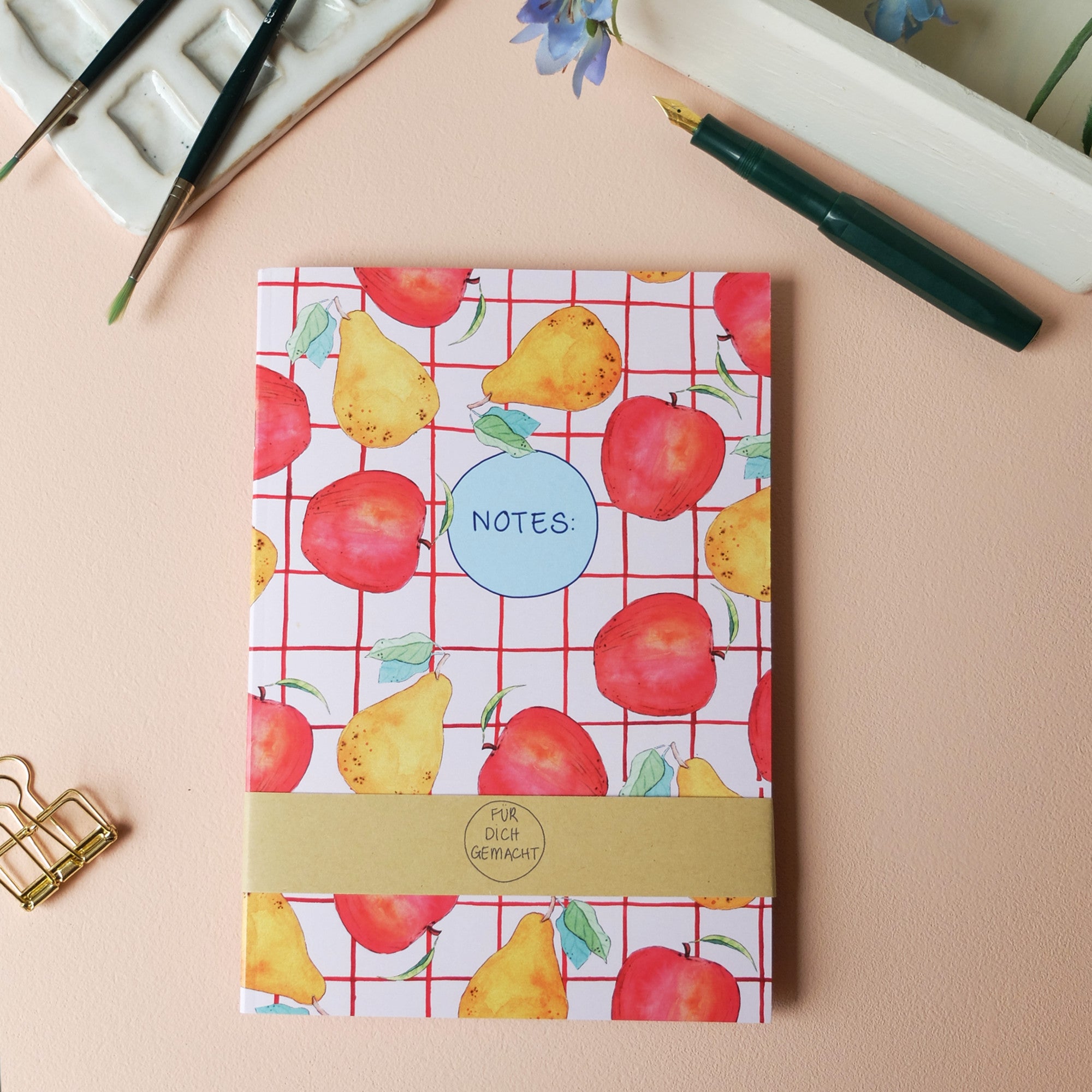 Notizbuch mit Obstmotiv, grüner Stift, Pinsel und eine Büroklammer liegen auf einem pfirsichfarbenen Untergrund.