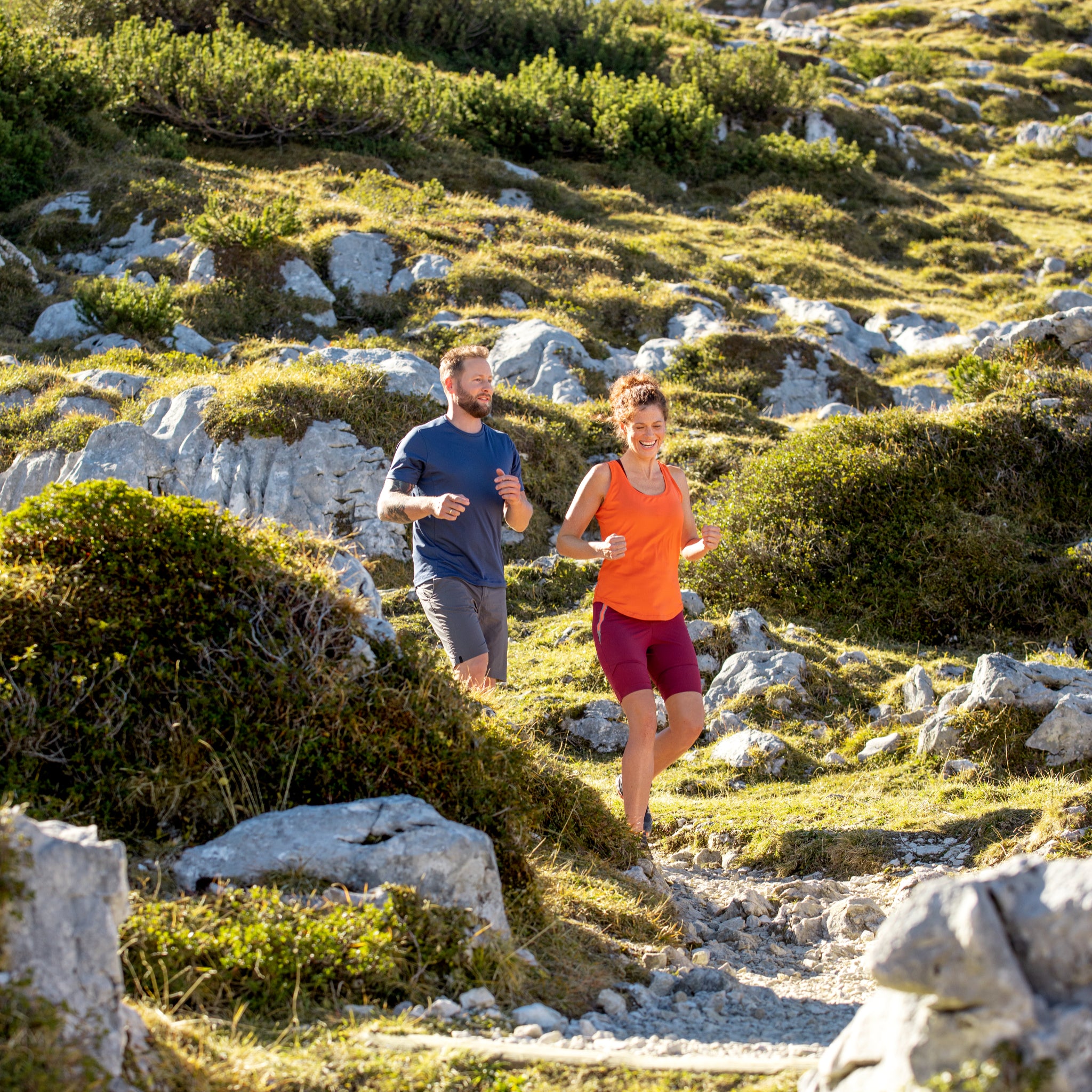 Ein Mann und eine Frau in sportlicher Kleidung laufen gemeinsam auf einem felsigen Bergpfad, umgeben von grünen Büschen und Sonnenlicht.