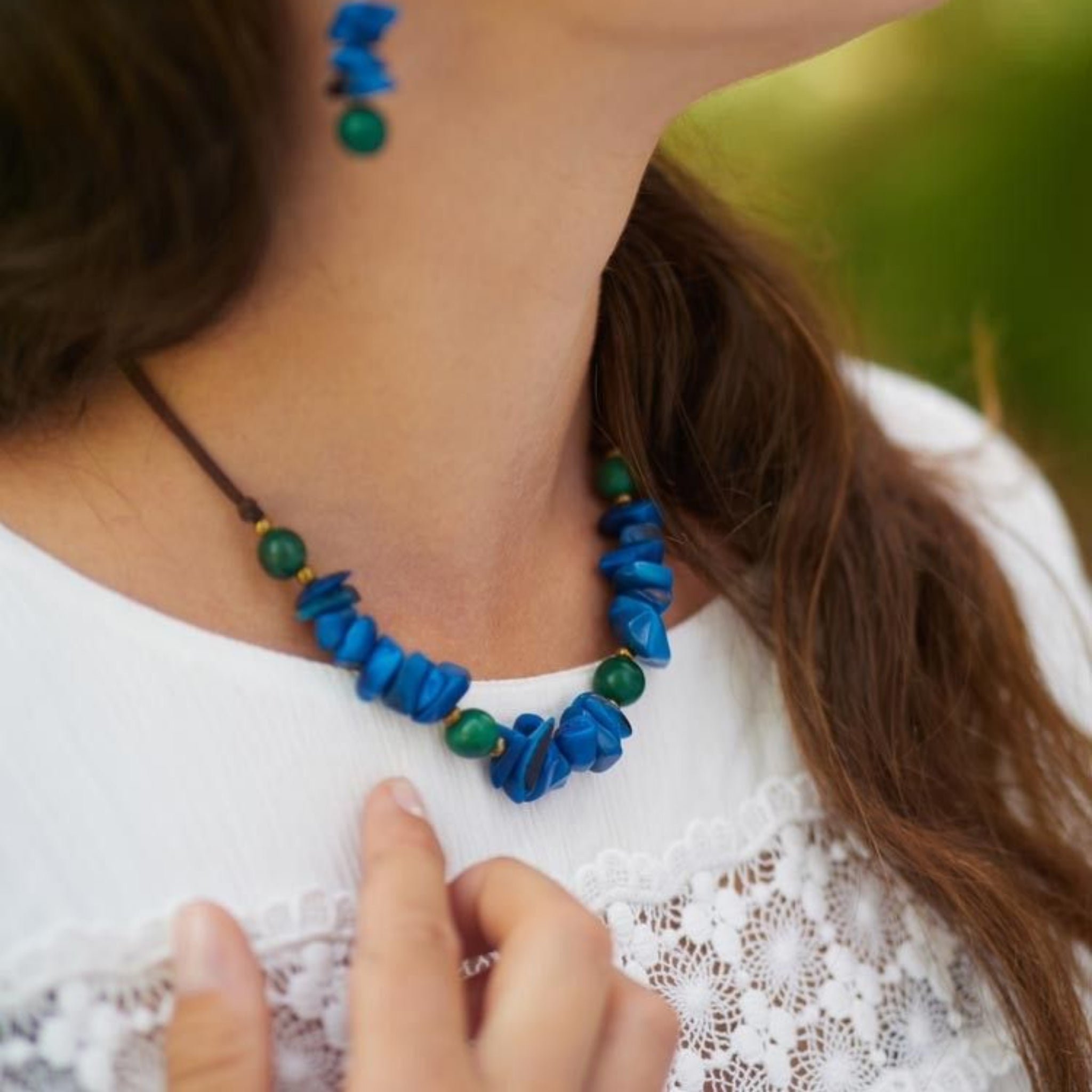 Eine Frau, die eine weiße Bluse mit Spitzenverzierung trägt, berührt eine Halskette aus blauen Steinen und grünen Perlen; sie trägt auch einen passenden blauen und grünen Ohrring. Ihr braunes Haar ist teilweise sichtbar.