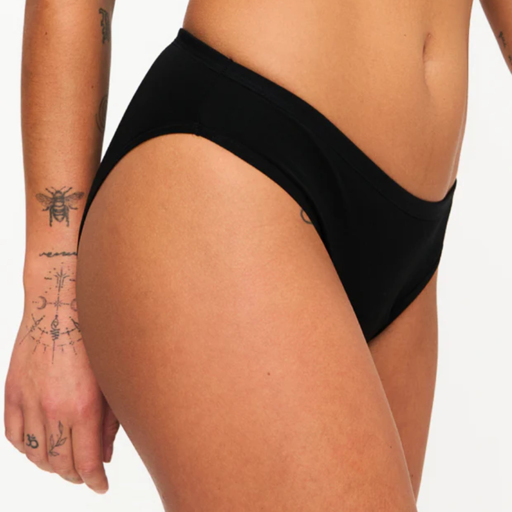 Eine Person, die eine schwarze Bikinihose trägt, die ihren Mittelteil und ihre Oberschenkel zeigt. Ihre Haut ist gebräunt und sie hat mehrere kleine Tätowierungen auf ihrem Arm und ihrer Hand. Der Hintergrund ist einfarbig weiß.