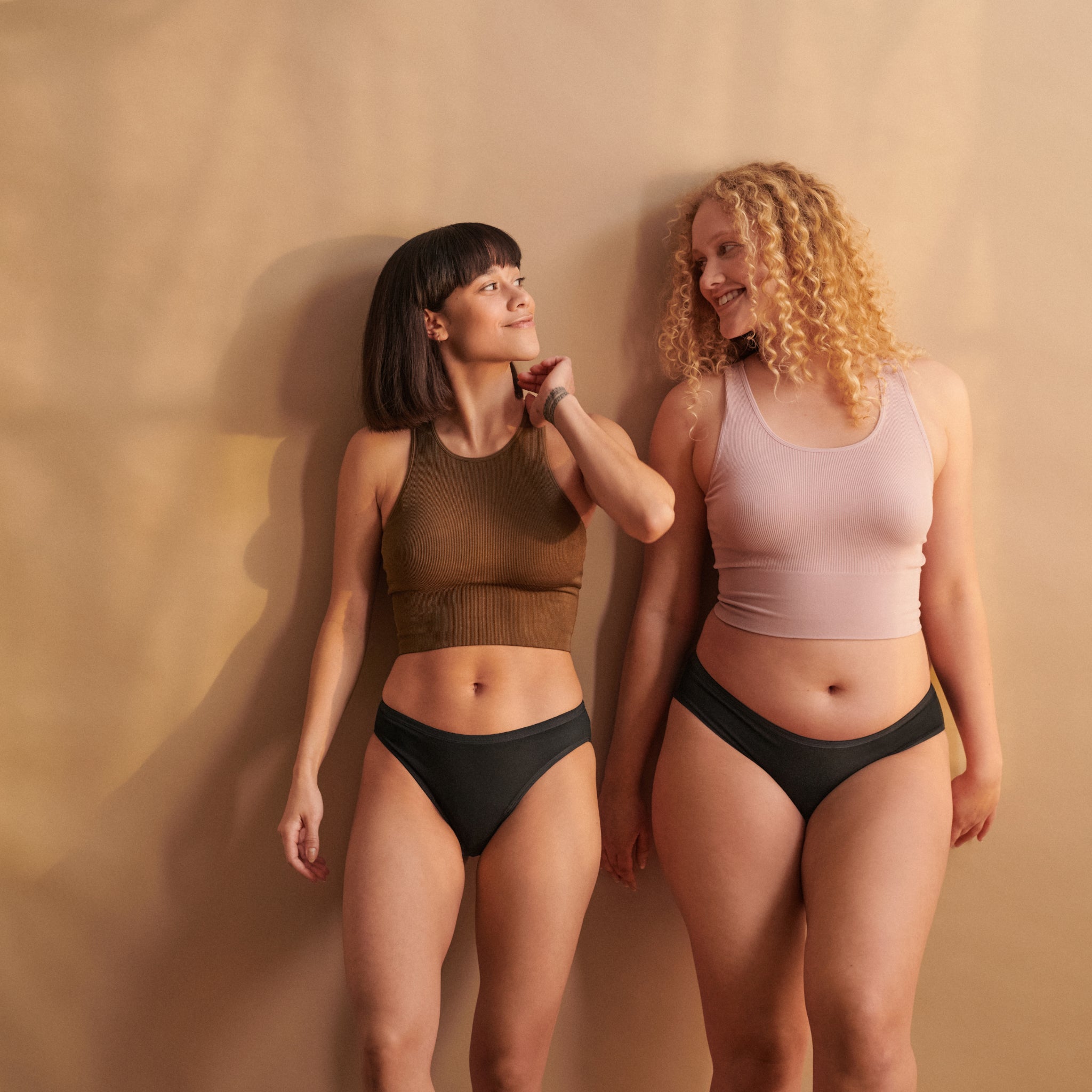 Zwei Frauen in Unterwäsche und ärmellosen Tops stehen lächelnd nebeneinander vor einem beigefarbenen Hintergrund.