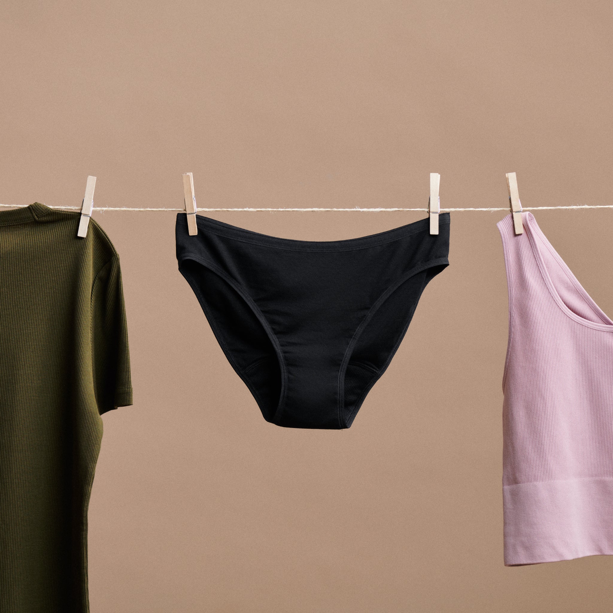 Dunkle Unterhose hängt auf einer Wäscheleine zwischen einem olivgrünen T-Shirt und einem rosafarbenen Trägertop.