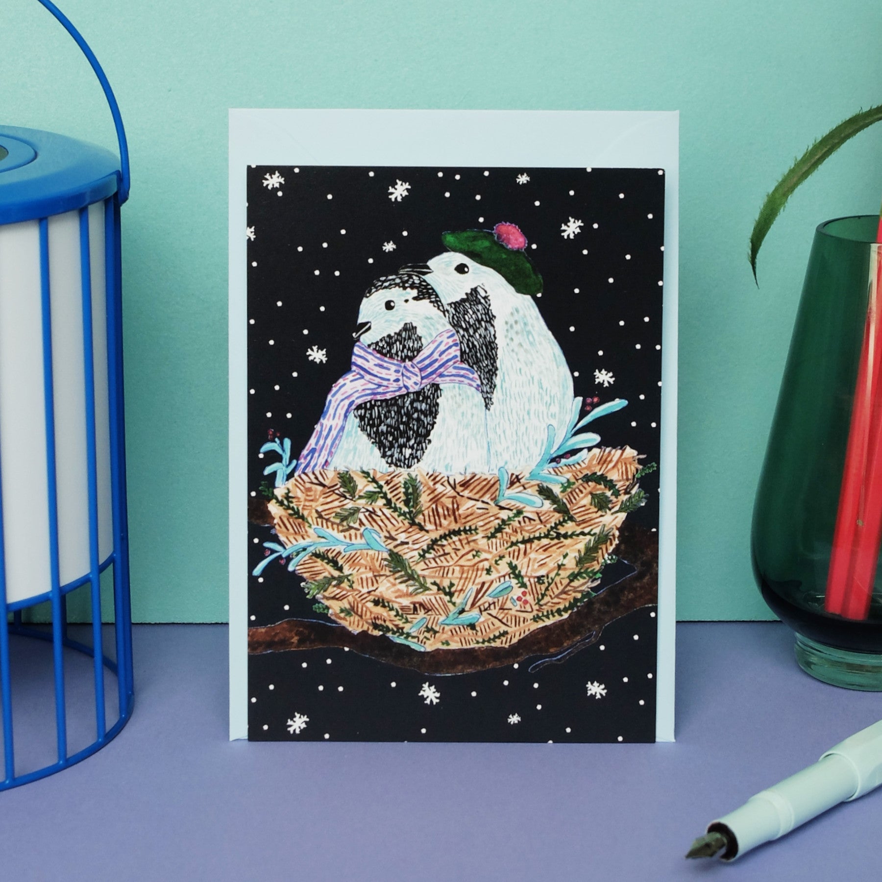 Eine Grußkarte zeigt zwei Pinguine mit Schals und Mützen, die in einem Nest mit verschneiten Zweigen kuscheln, vor einem schwarzen Hintergrund mit fallenden Schneeflocken. Die Karte steht auf einer lila Fläche.
