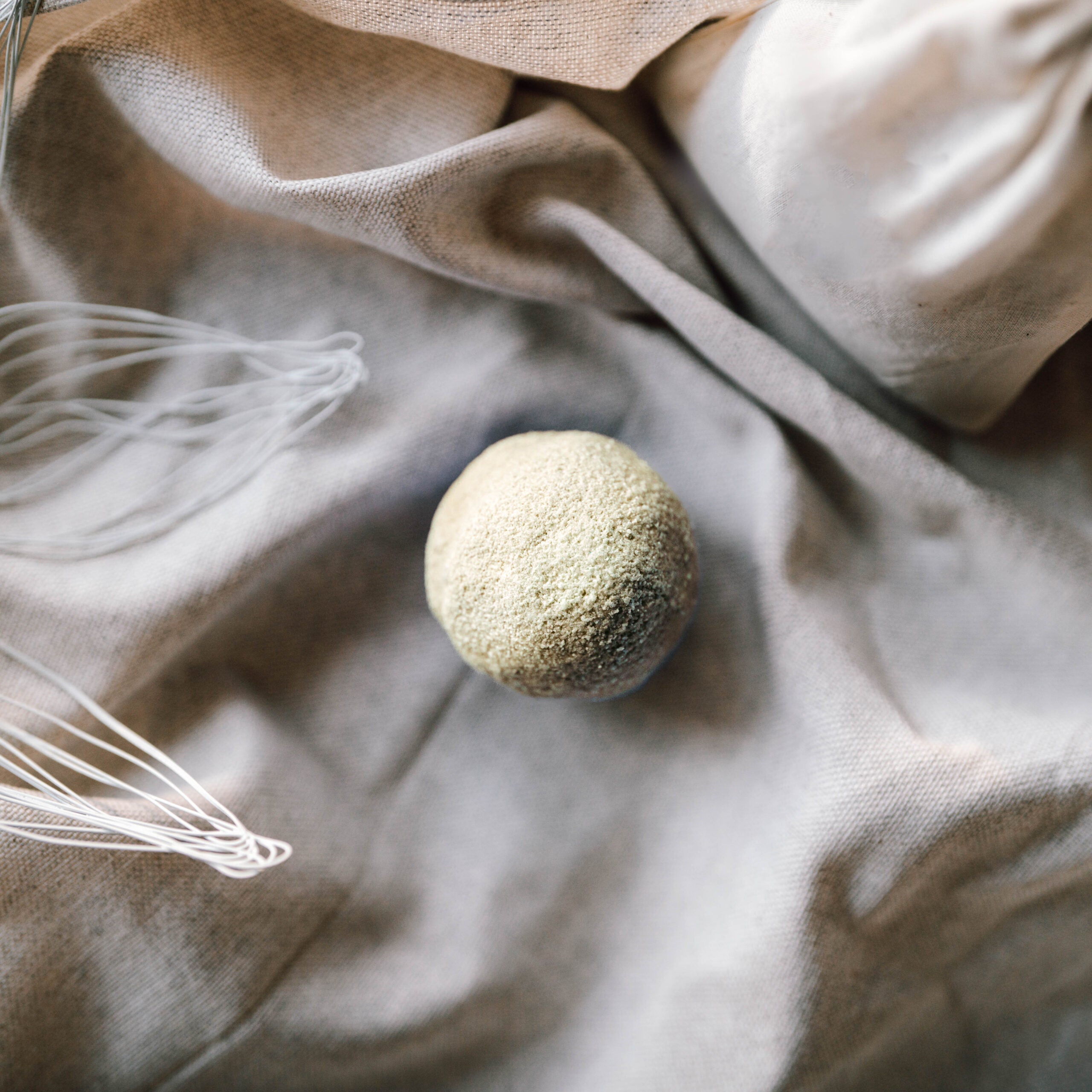 Eine runde, mit Mehl bestäubte Teigkugel liegt auf einem beigen Tuch, daneben stehen ein Schneebesen und ein mit einem Tuch bedeckter Gegenstand, die eine rustikale Backszene darstellen.