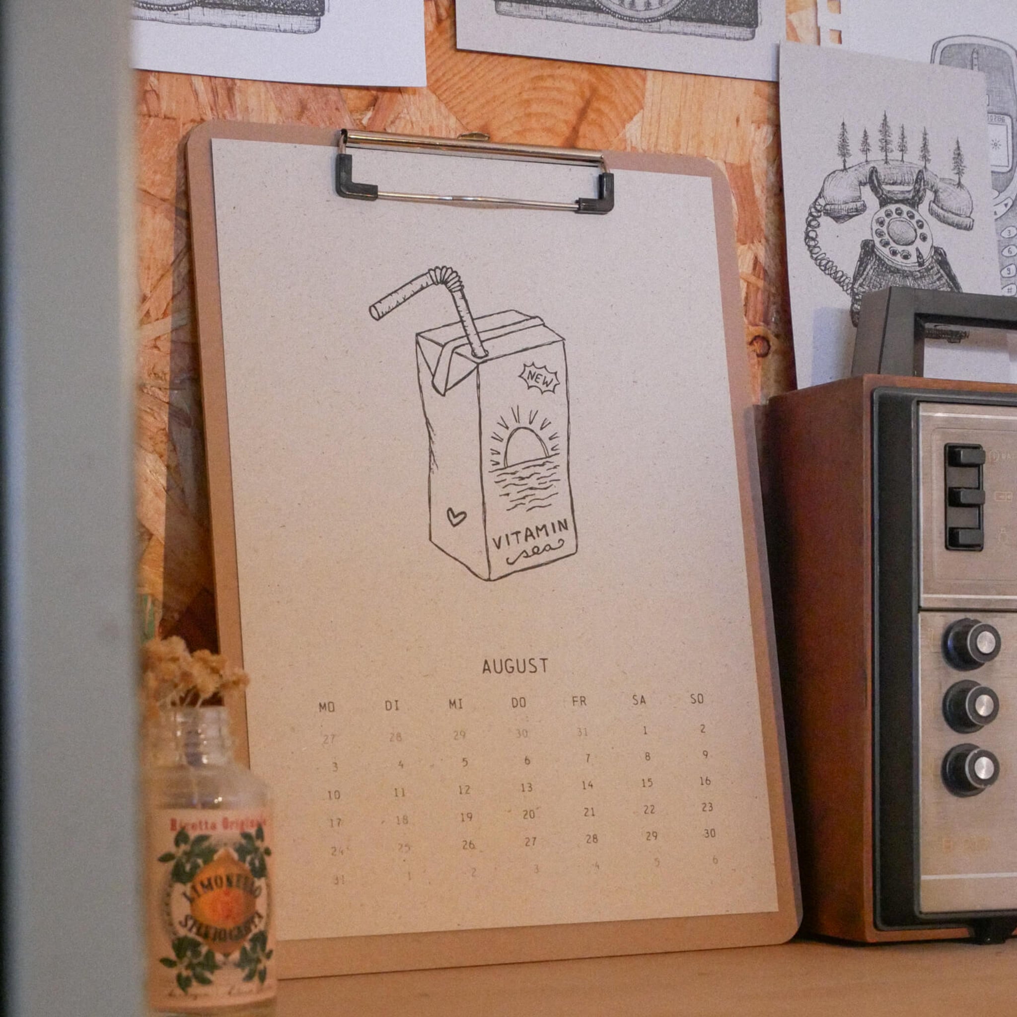 Ein Klemmbrett mit einem Kalenderblatt für den Monat August, auf dem ein handgezeichneter Saftkarton mit der Aufschrift "Vitamin" zu sehen ist, liegt auf einem Schreibtisch neben einem alten Radio und einer kleinen dekorativen Flasche, hinter der Skizzen an die Wand gepinnt sind.