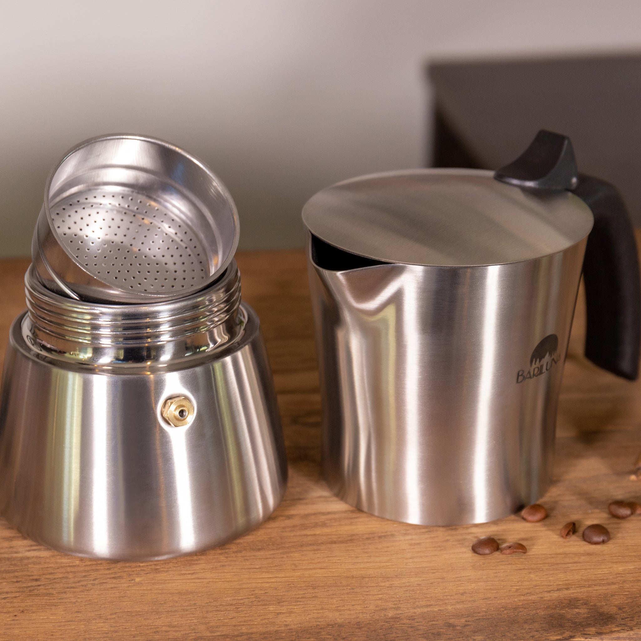 Edelstahl-Espressokocher, geöffnet, auf Holztisch, mit einem Siebeinsatz und einigen verstreuten Kaffeebohnen daneben.
