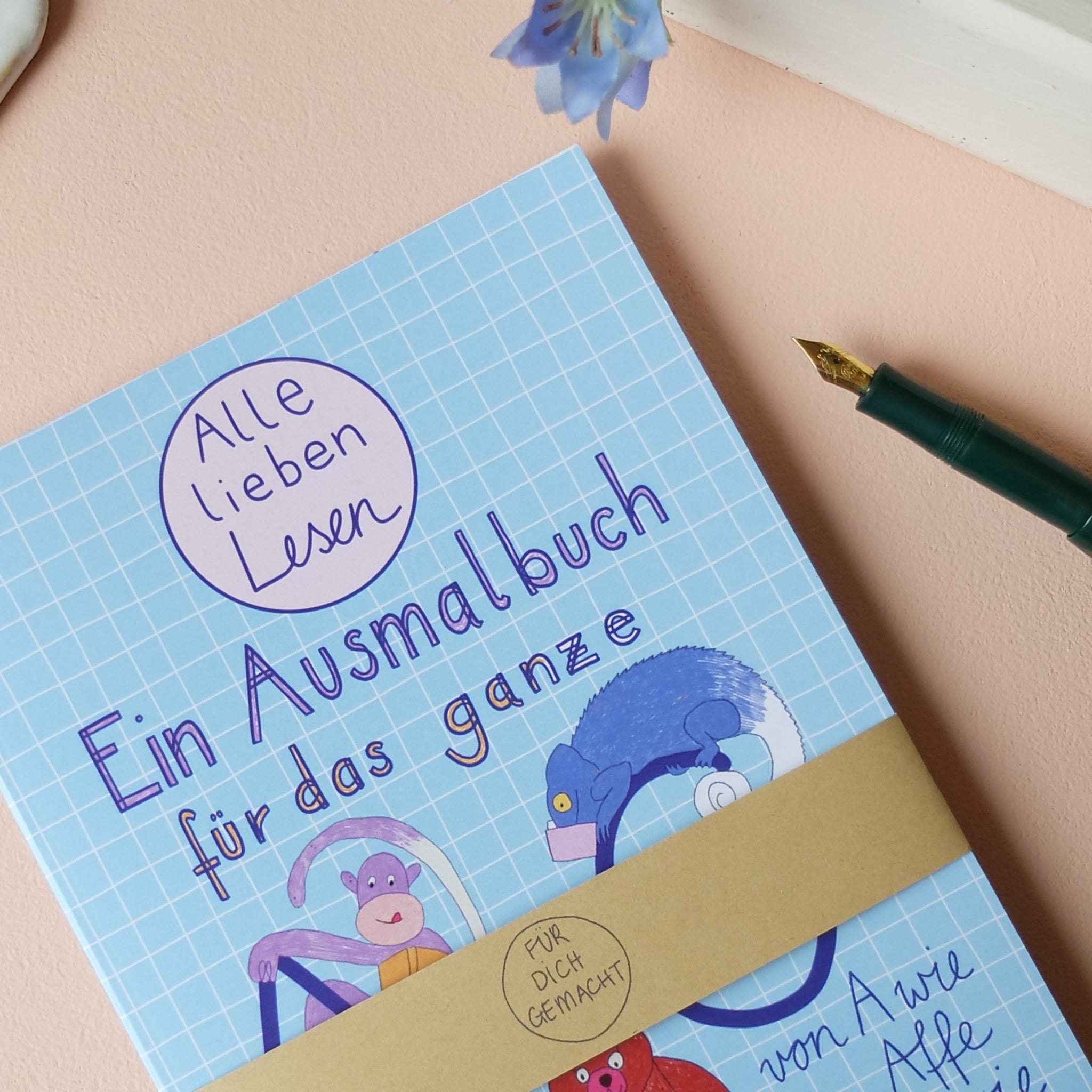 Ein Ausmalbuch mit Illustrationen von Tieren auf einem blauen, karierten Hintergrund, daneben ein grüner Füllfederhalter.