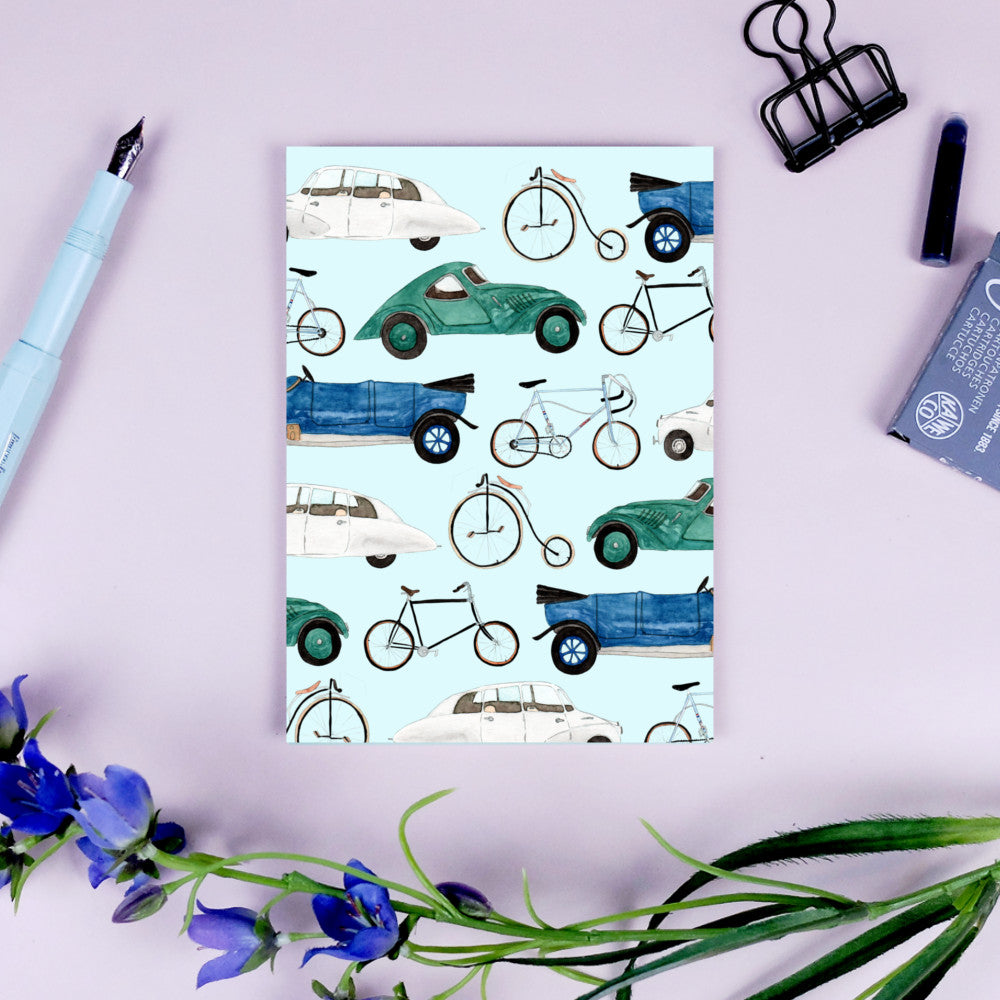 Postkarte mit Illustrationen von Autos und Fahrrädern auf hellblauem Hintergrund, daneben Stift und Blumen.