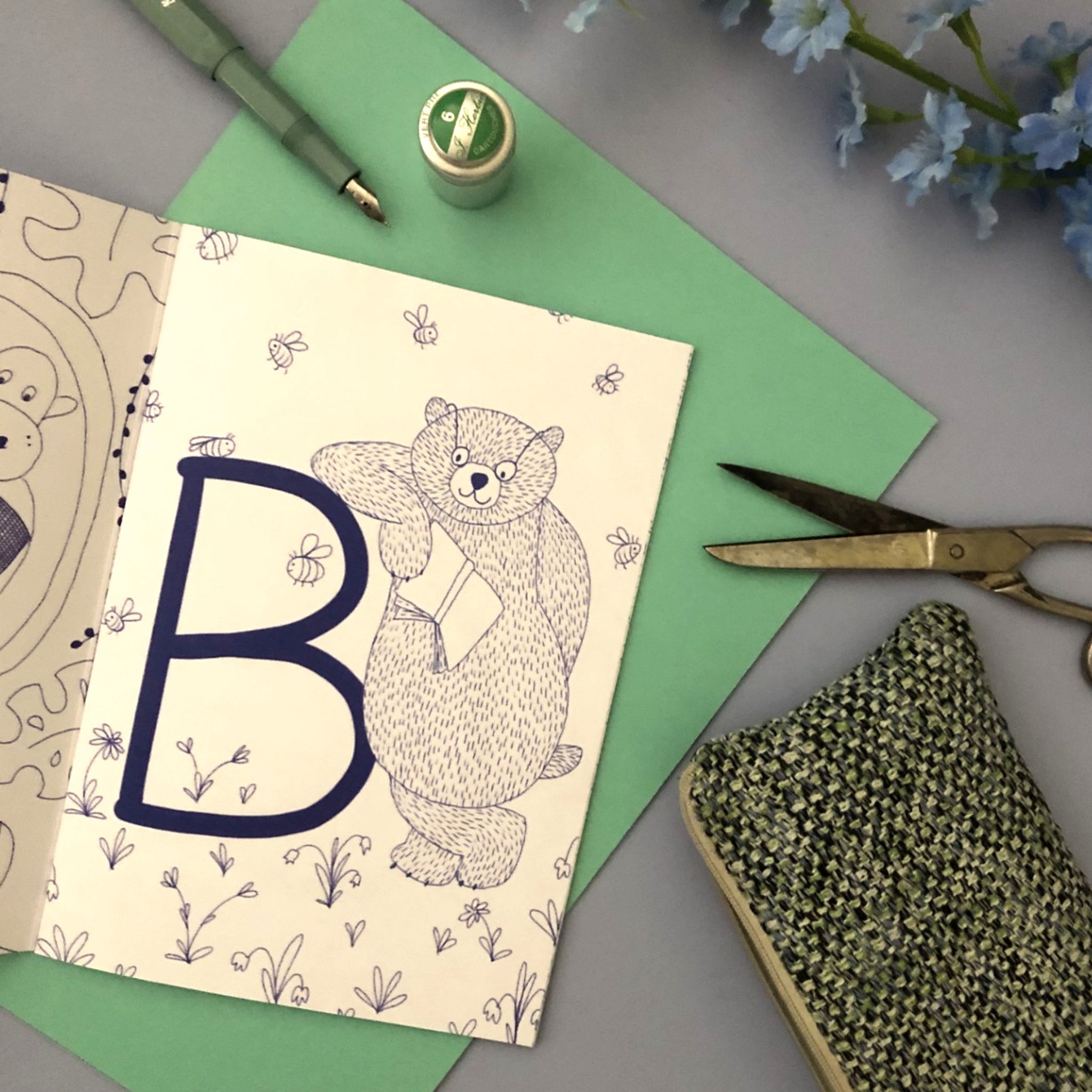 Ein gezeichneter Bär mit einem Buch umgeben von Bienen auf einer Karte, umgeben von Stift, Schere und Garn.