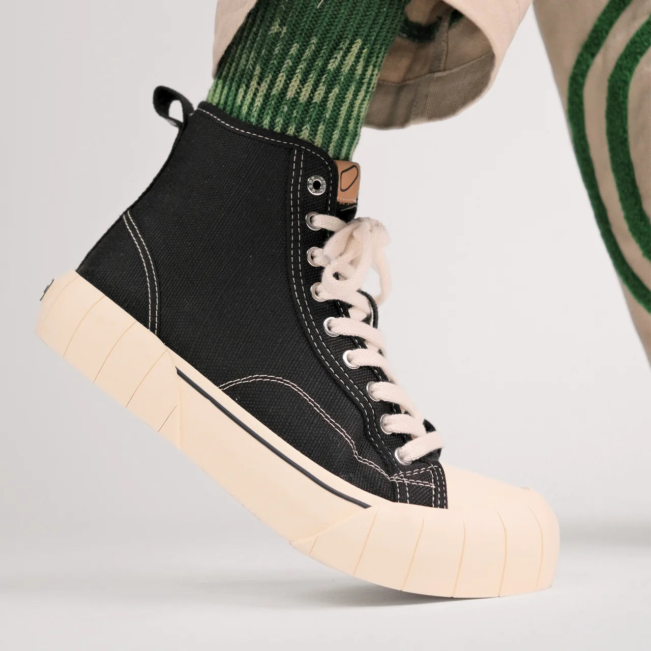 Schwarz-weißer High-Top-Sneaker an einem Fuß mit grün gestreiften Socken und beigefarbener Hose auf grauem Hintergrund.