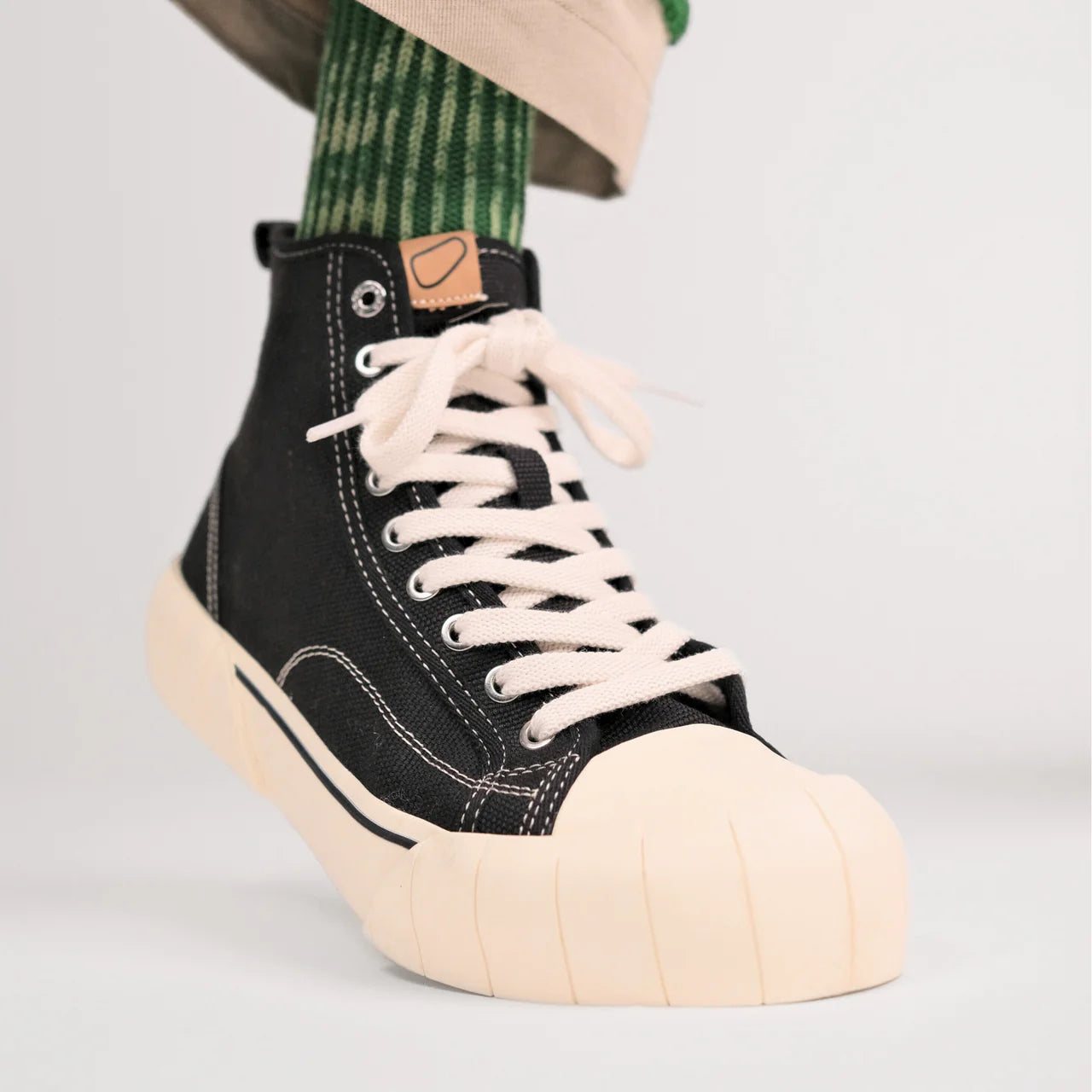 Schwarzer High-Top-Sneaker mit weißer Sohle und Schnürsenkeln, getragen mit grünen Socken und beiger Hose.