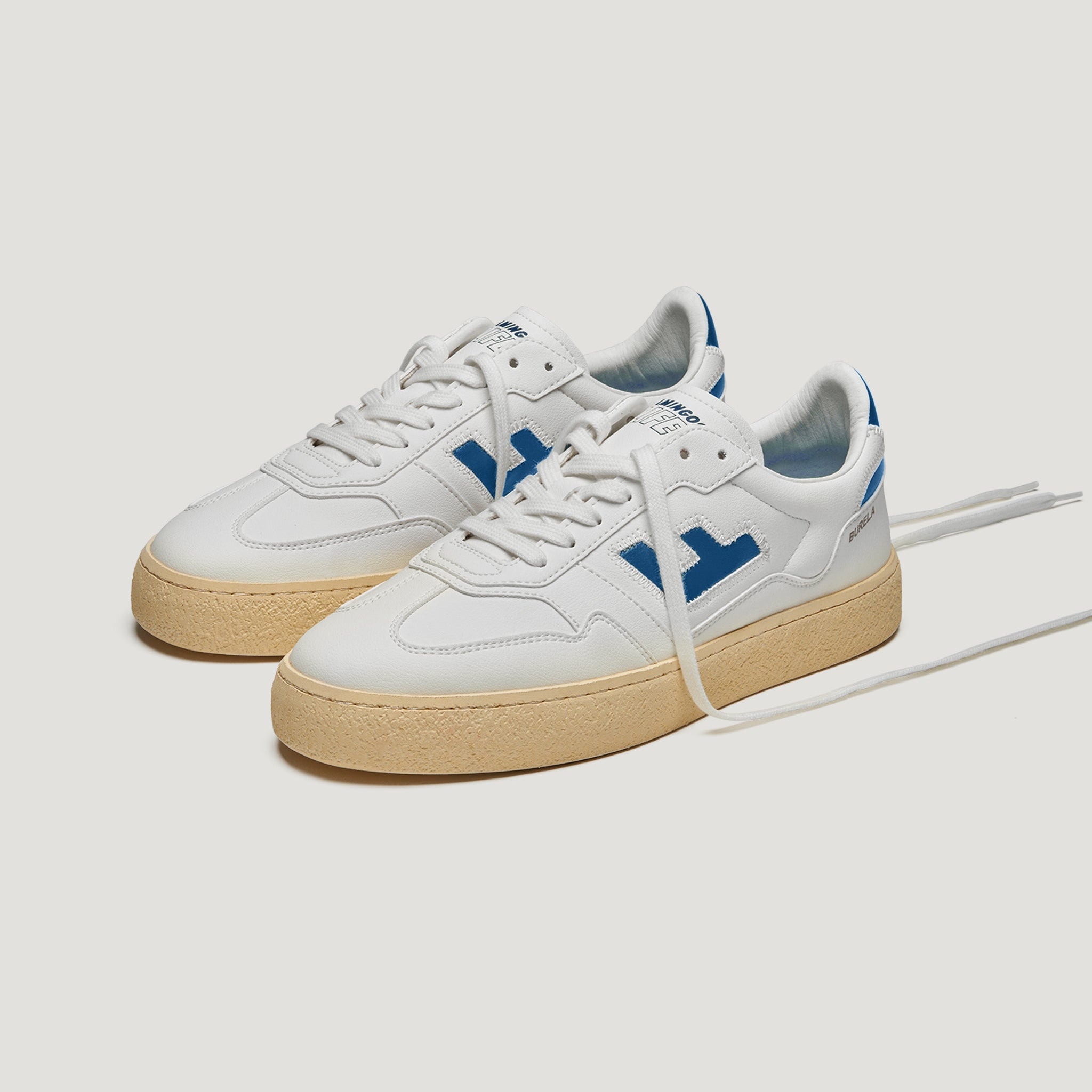 Ein Paar weiße Turnschuhe mit beiger Gummisohle und blauen Akzenten, darunter blaue Logos an den Seiten und blaue Details an der Ferse. Die Schnürsenkel sind weiß und ein Schnürsenkel ist gelöst.