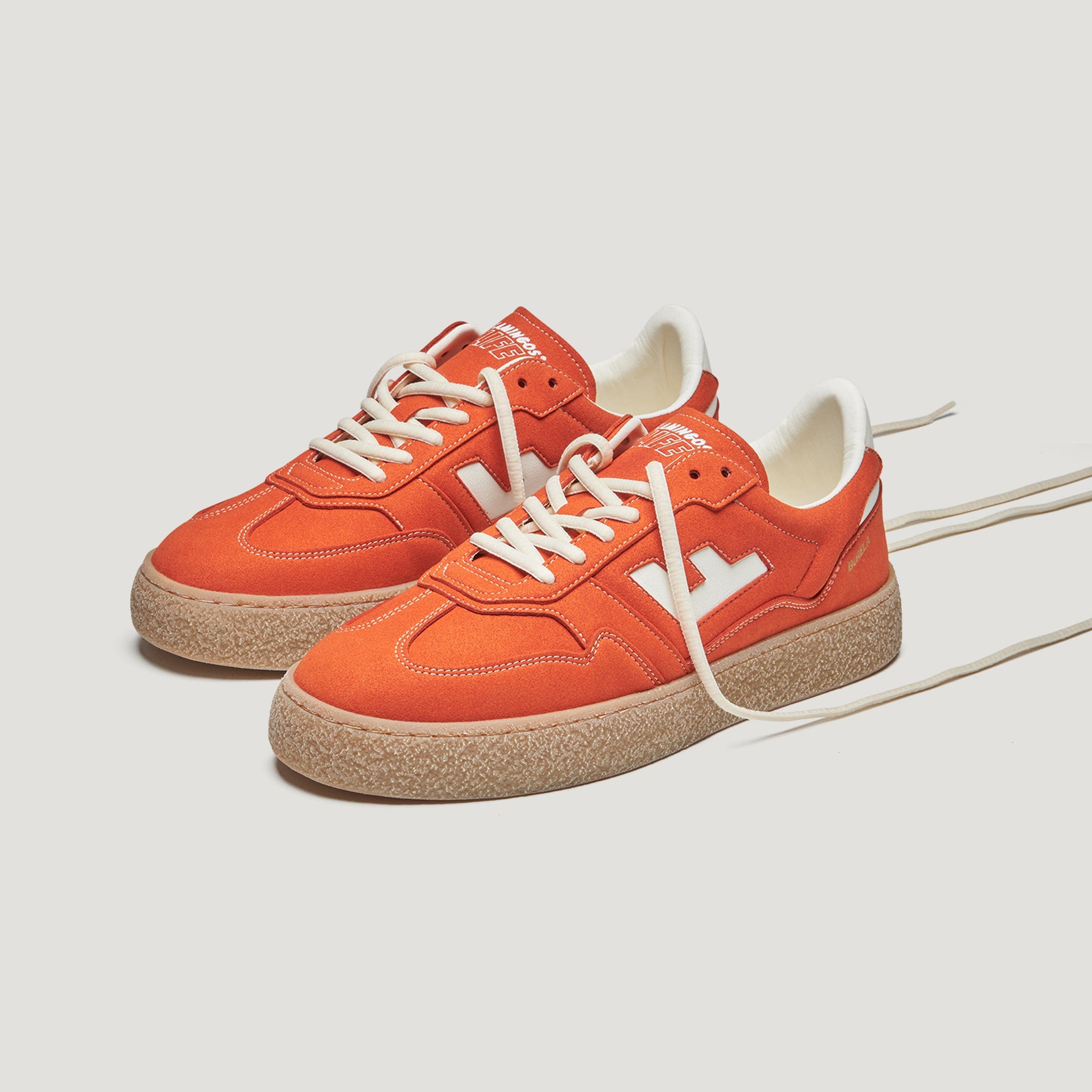 Ein Paar orangefarbene Wildleder-Sneakers mit weißen Schnürsenkeln, weißen Akzenten und beigen, strukturierten Gummisohlen auf einem neutralen Hintergrund.