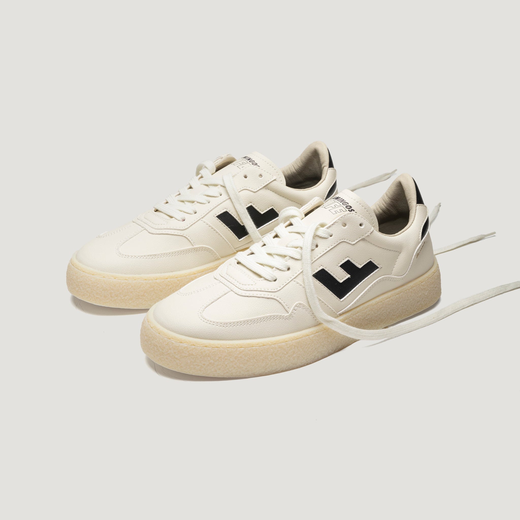 Ein Paar cremefarbene Turnschuhe mit beiger Sohle, schwarzem Logo an den Seiten und weißen Schnürsenkeln vor einem schlichten hellen Hintergrund.