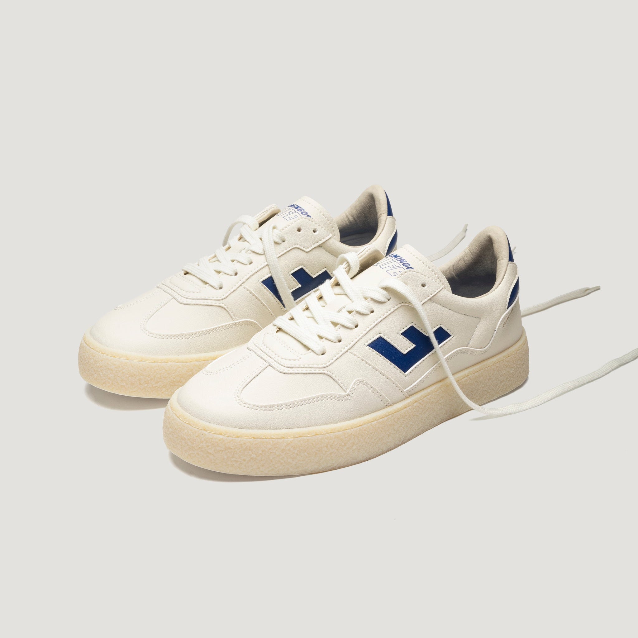 Ein Paar weiße Turnschuhe mit beigen Sohlen, blauen Akzenten und einem auffälligen blauen L-Muster an den Seiten auf einem hellgrauen Hintergrund. Ein Schnürsenkel ist lose und nach außen verlängert.