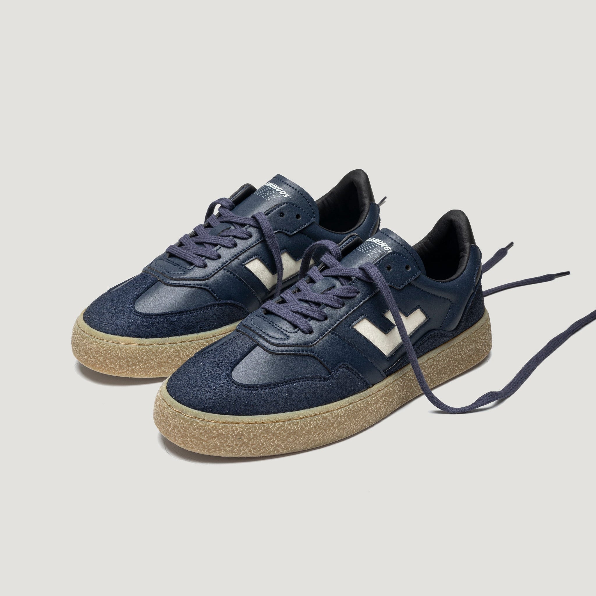 Ein Paar marineblaue Turnschuhe mit beigen, strukturierten Sohlen, weißen Akzenten an den Seiten und offenen Schnürsenkeln vor einem einfarbig hellgrauen Hintergrund.