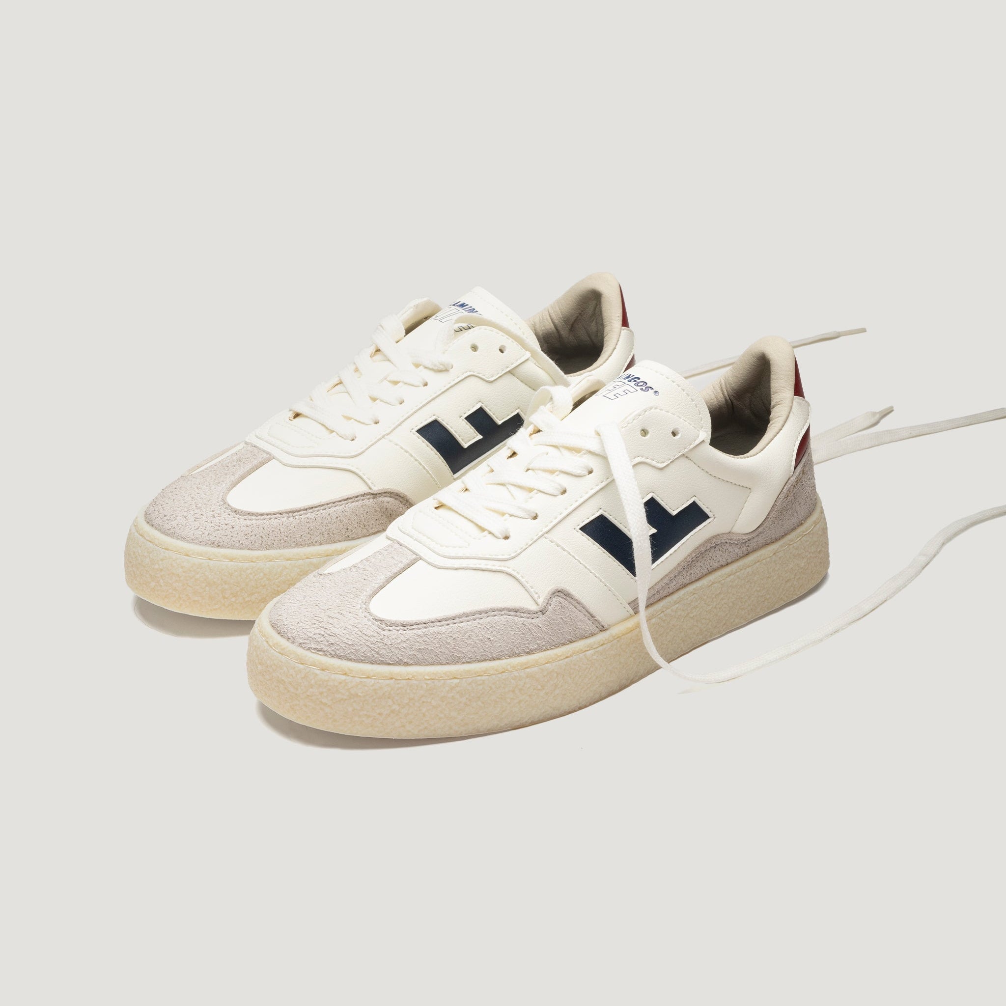 Ein Paar stylische Low-Top-Sneaker mit beiger Sohle, cremefarbenem Obermaterial, grauen Wildlederkappen, marineblauen Seitendetails und weißen Schnürsenkeln vor einem schlichten, hellen Hintergrund.