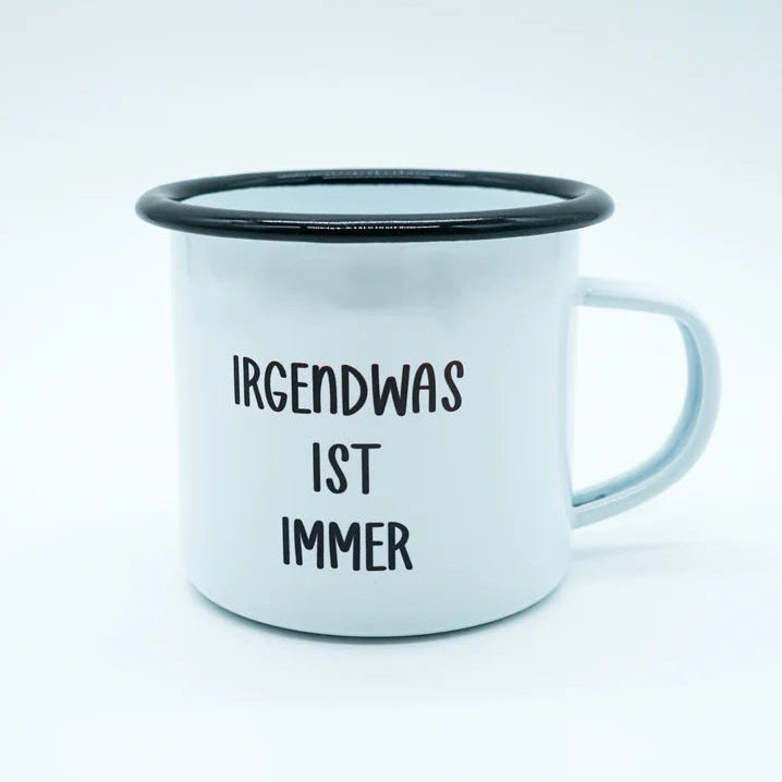 Weißer Emaillebecher mit schwarzem Rand und dem Schriftzug IRGENDWAS IST IMMER in schwarzer Schrift auf der Vorderseite vor einem schlichten weißen Hintergrund.