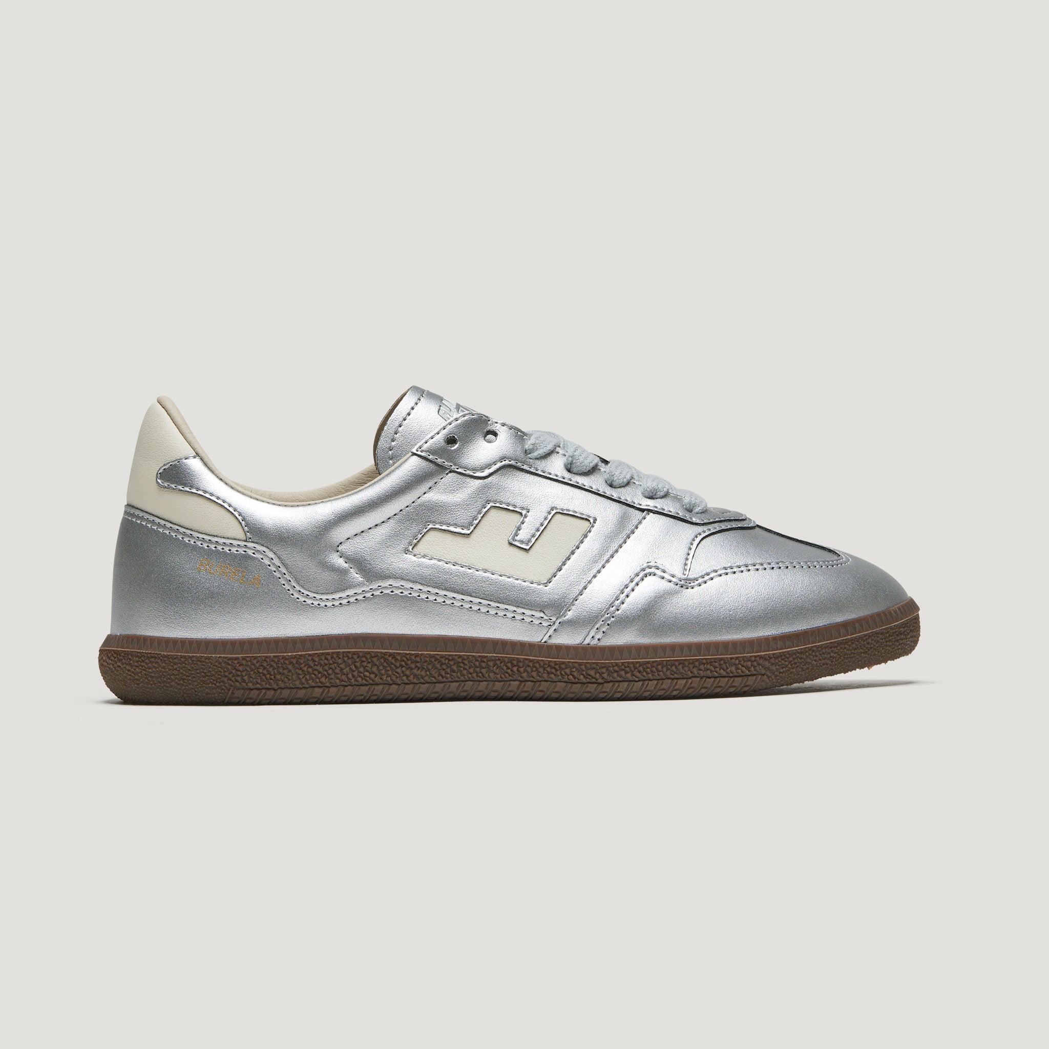 Silberner Sneaker mit brauner Sohle, seitlicher Logoapplikation und weißen Details auf grauem Hintergrund.