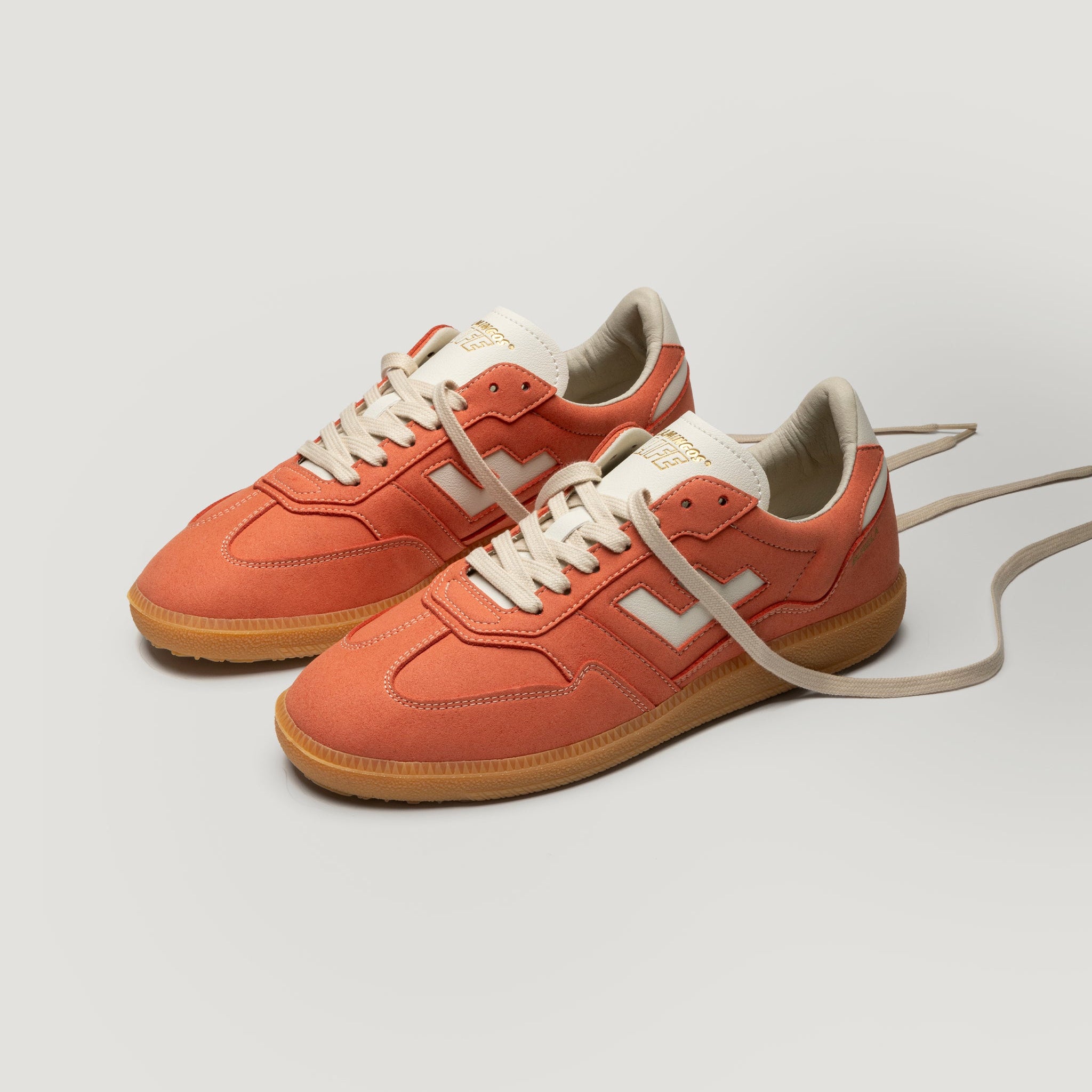 Ein Paar orangefarbene Turnschuhe mit weißen Schnürsenkeln, weißen seitlichen Details und hellbraunen Gummisohlen sind auf einem einfarbig weißen Hintergrund abgebildet; der Schnürsenkel des einen Schuhs ist locker nach außen verlängert.