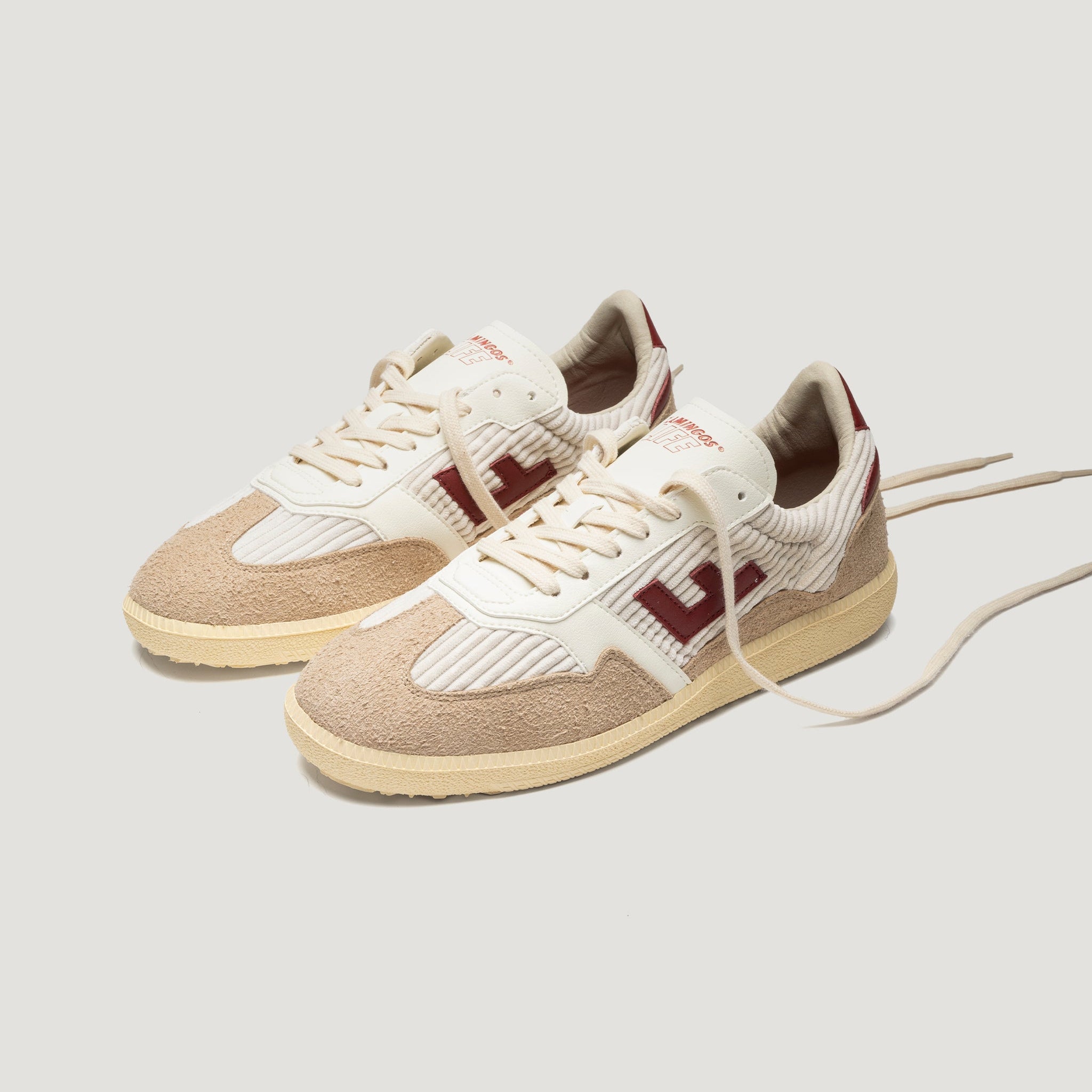 Ein Paar beige- und cremefarbene Turnschuhe mit strukturierten Wildledereinsätzen, weißen Schnürsenkeln und kastanienbraunen Akzenten, darunter ein großes H-Logo an der Seite, auf hellem Hintergrund.